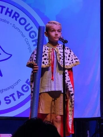 A young girl dressed in a costume with a crown and cape, standing on stage in front of a microphone and holding a long staff, during a school play or performance.