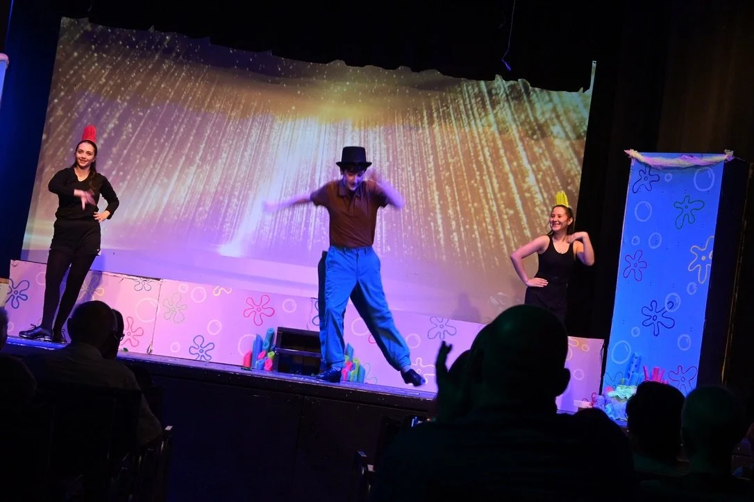 Three performers on stage with colorful children's-themed backdrop, including a woman on the left, a man in the middle wearing a black hat, and a woman on the right, engaging in a theatrical or dance performance.
