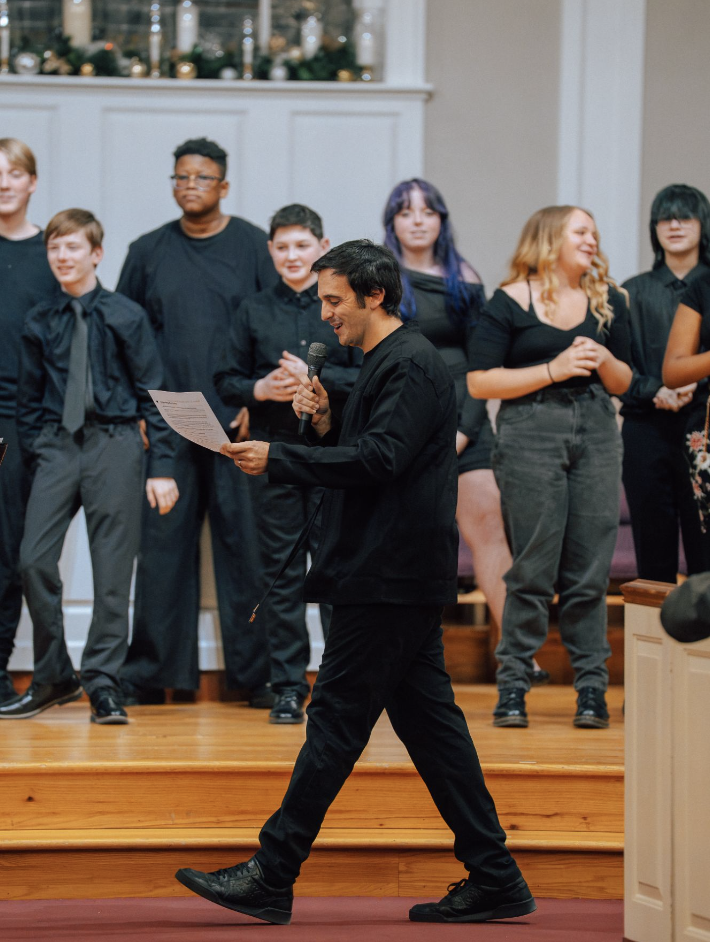 A smiling man with dark hair, wearing a white t-shirt, an unbuttoned blue plaid shirt, and jeans, standing outdoors in a natural setting with trees and grass.