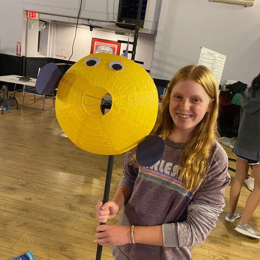 A girl holding a yellow paper lantern with googly eyes, black paper ears, and black paper paws, standing indoors with chairs and tables in the background.