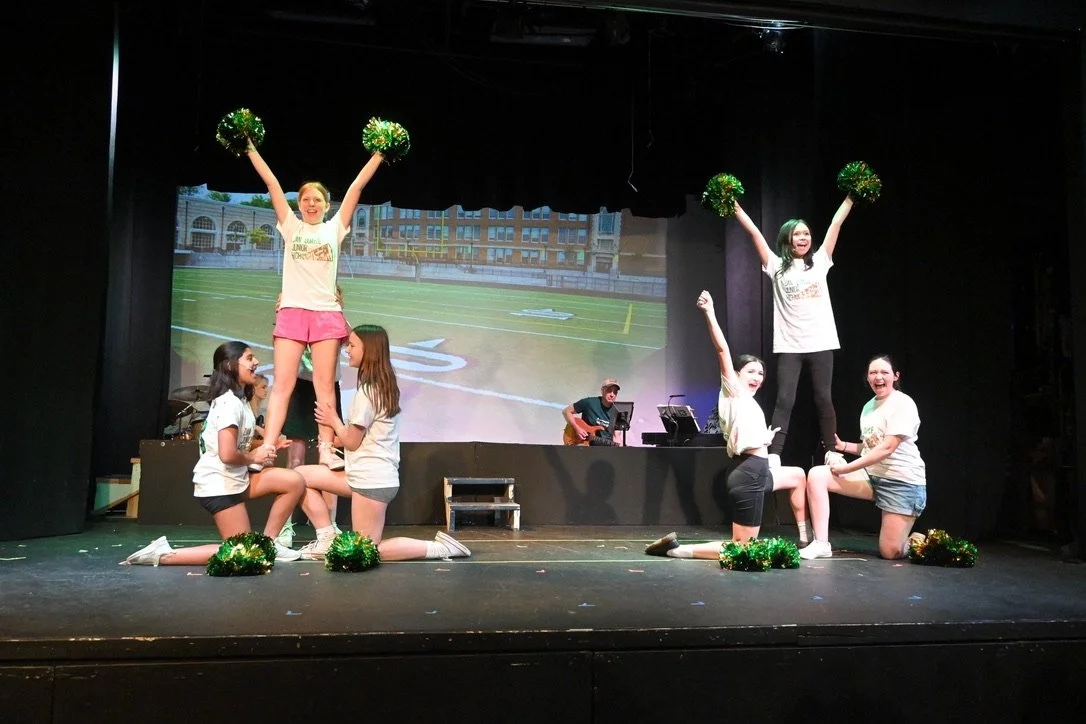 Cheerleading routine on stage with seven girls, two in the center lifted by others, holding green pom-poms, with a musician playing guitar in the background.