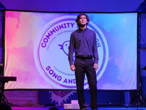 A young man stands on a stage in front of a large screen displaying a logo for a community youth musical organization. The stage is lit with colorful lighting.
