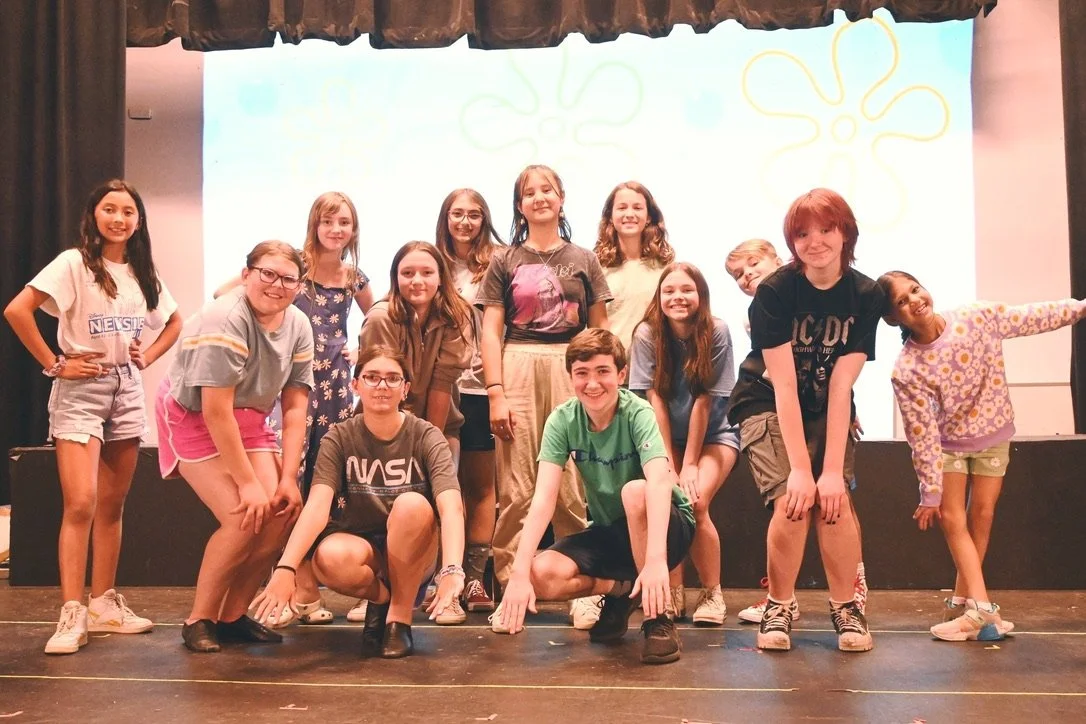 Group of children and teenagers posing on stage with a screen displaying Jack-O-Lanterns or pumpkin drawings in the background.