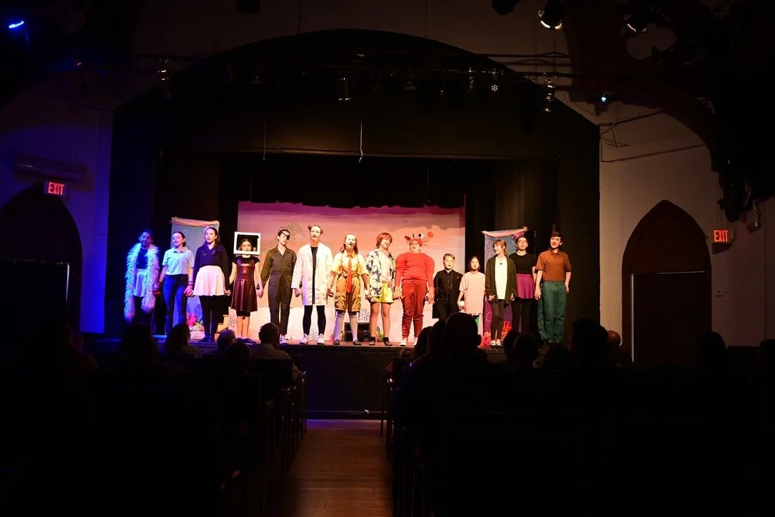 A group of children and teenagers standing on a stage during a theatrical performance, with a colorful backdrop and stage lighting.
