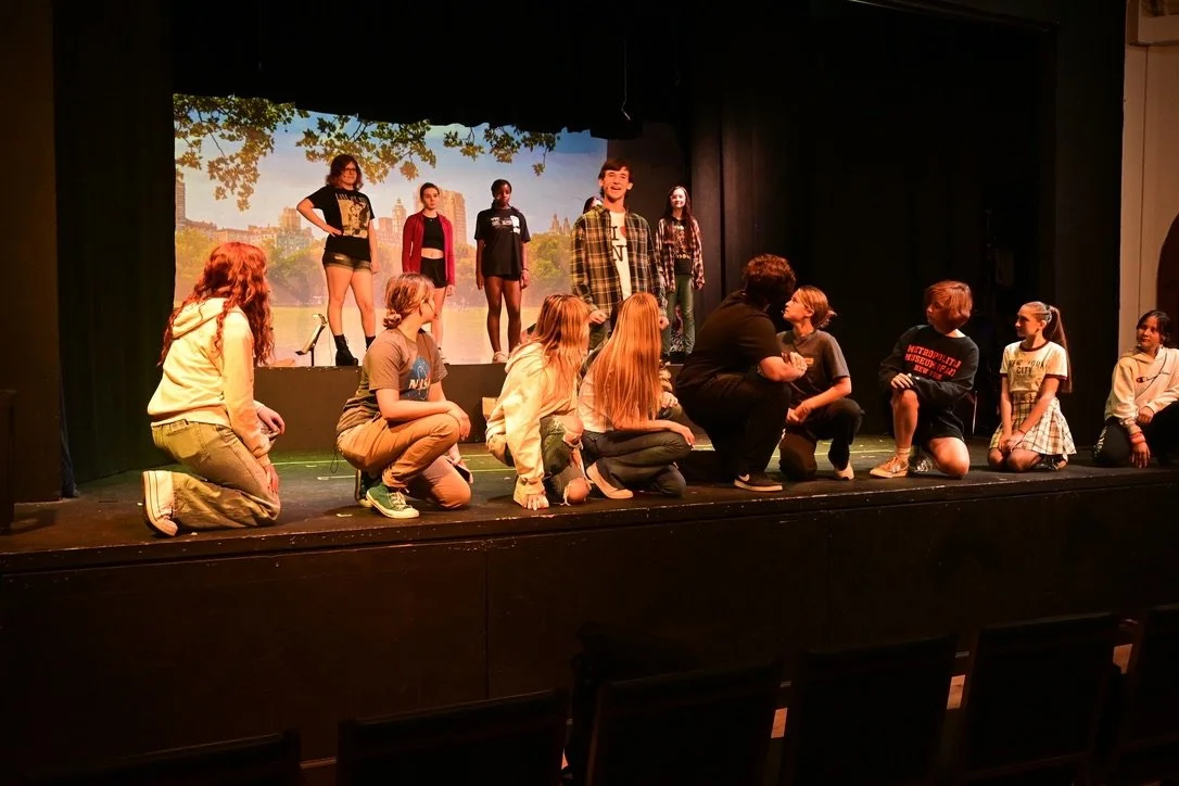 A group of children and teens on a theater stage, practicing or performing a play, with some kneeling and others standing against a backdrop of a park scene.