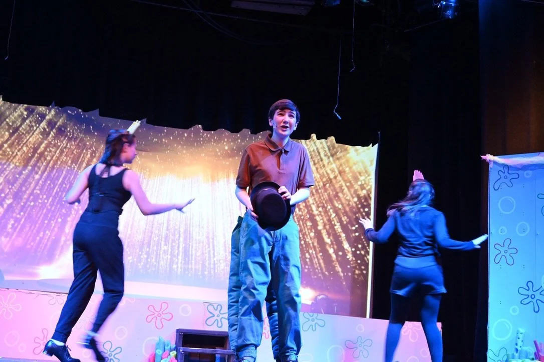 Three people performing on stage with colorful backdrops and props, engaged in a theatrical scene.