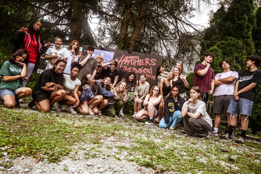 A group of young people posing outdoors in front of a sign for the musical 'Heathers' with trees and bushes in the background.