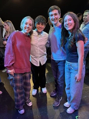 Group of four teenagers smiling and posing together at a social event, with others in the background.