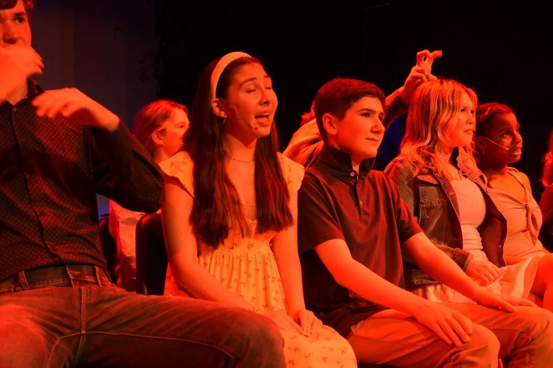 A group of young people sitting on stage, appearing to be engaged in a performance or rehearsal, illuminated by red stage lighting.