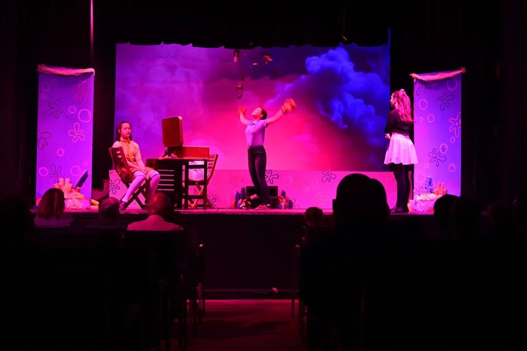 Three young women performing a play on a stage with colorful backdrop and decorations, audience watching.