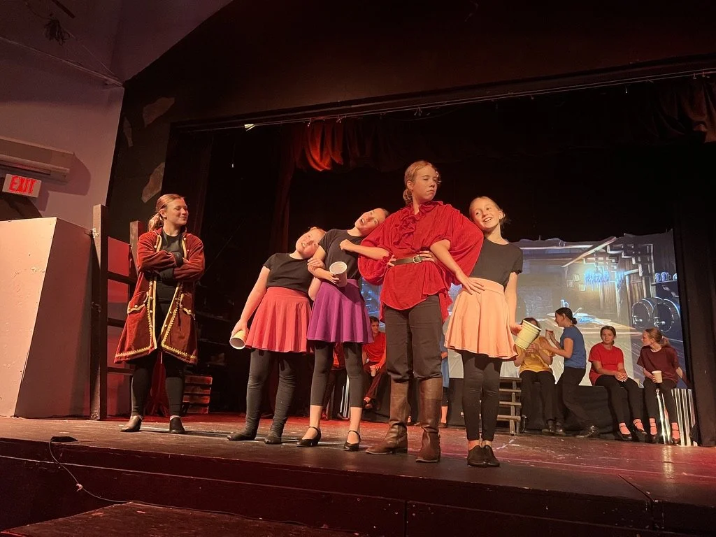 Children performing on stage in a theater, dressed in costumes, with some sitting and some standing, during a school play or musical.