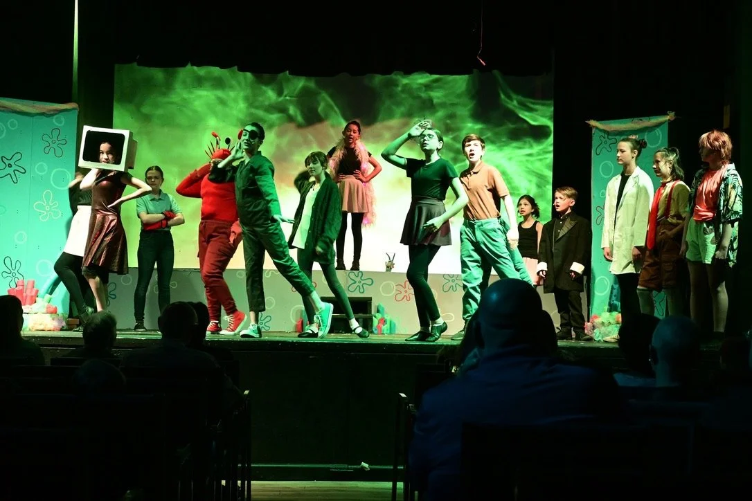 Children perform a stage play with various costumes, including a person with a square headpiece and a crab costume, against a green background, with audience members watching.