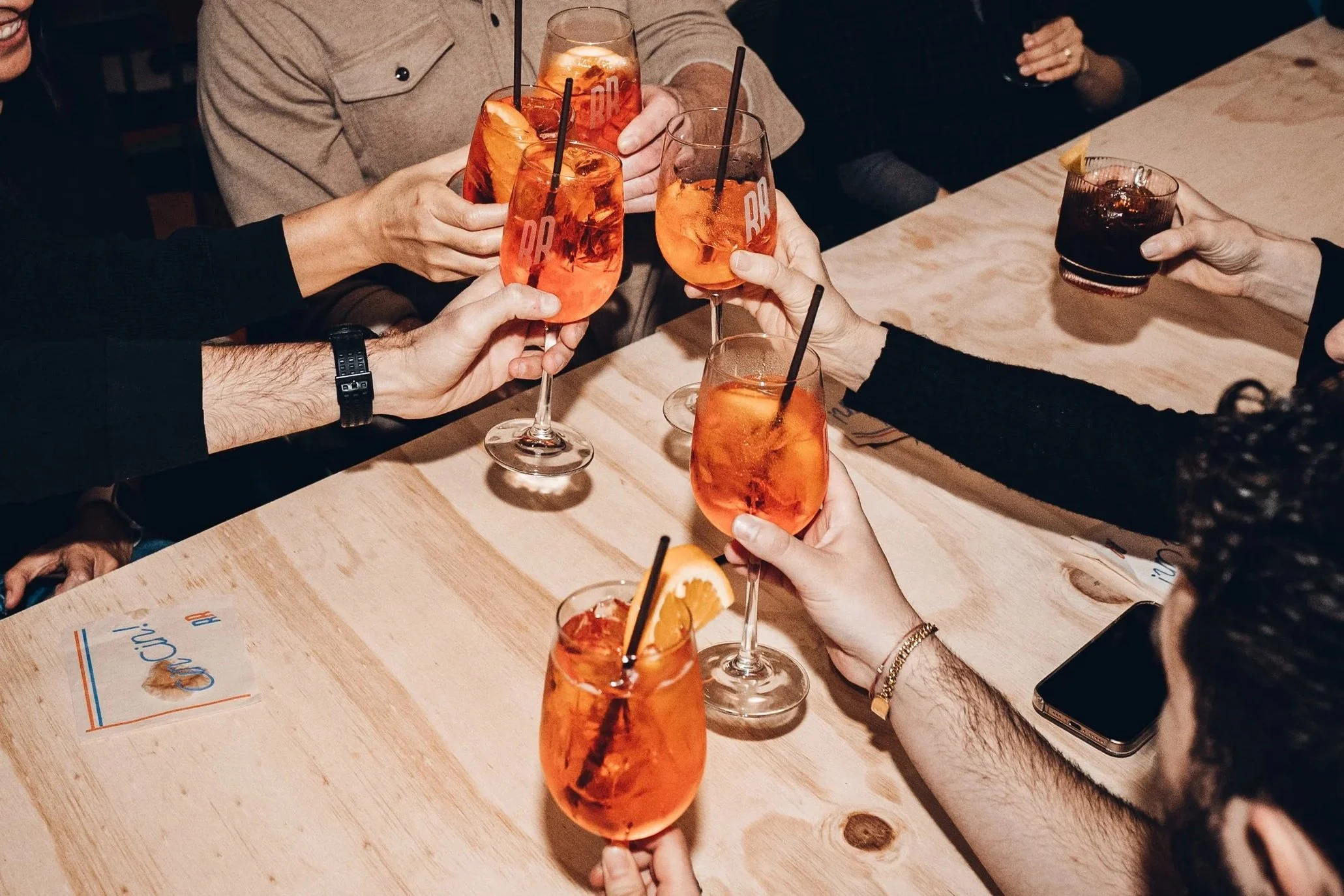 People clinking glasses filled with orange-colored drinks with ice and orange slices at a table in a social gathering.