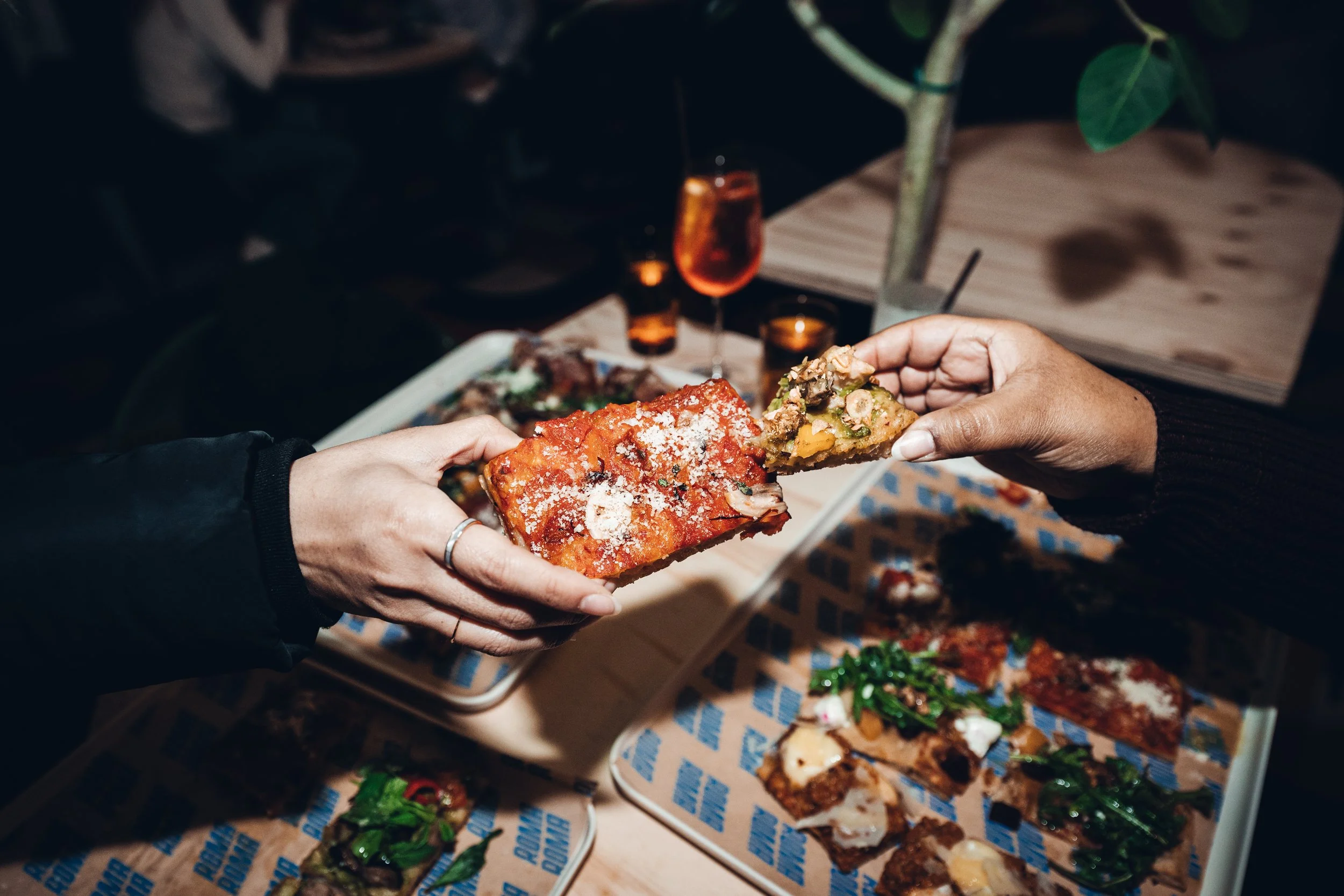 Hands holding a slice of pizza with tomato sauce and toppings at a party table with various food and drinks.
