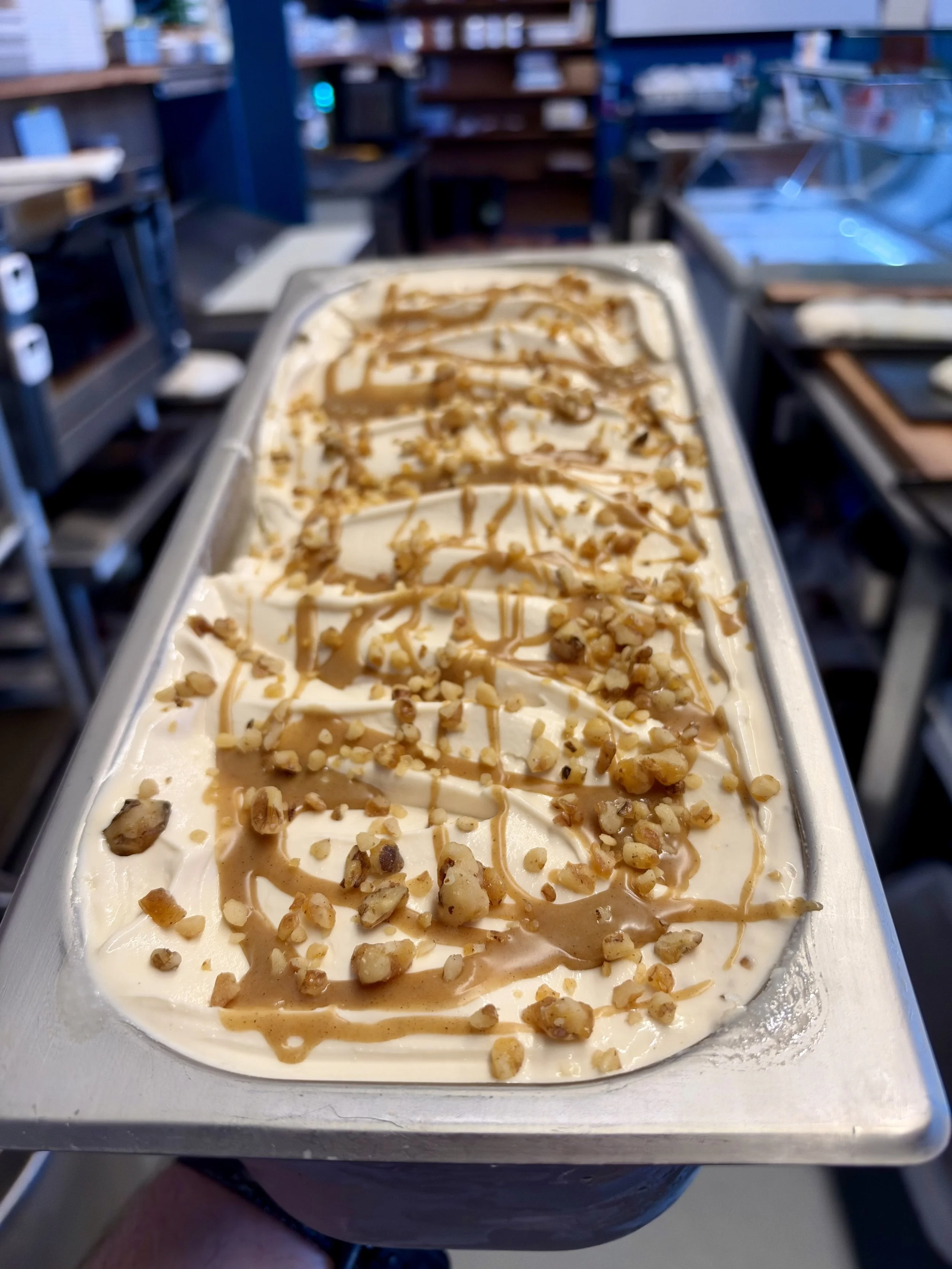 A tray of vanilla ice cream with caramel drizzle and chopped nuts on top, placed on a stainless steel counter in a commercial kitchen.