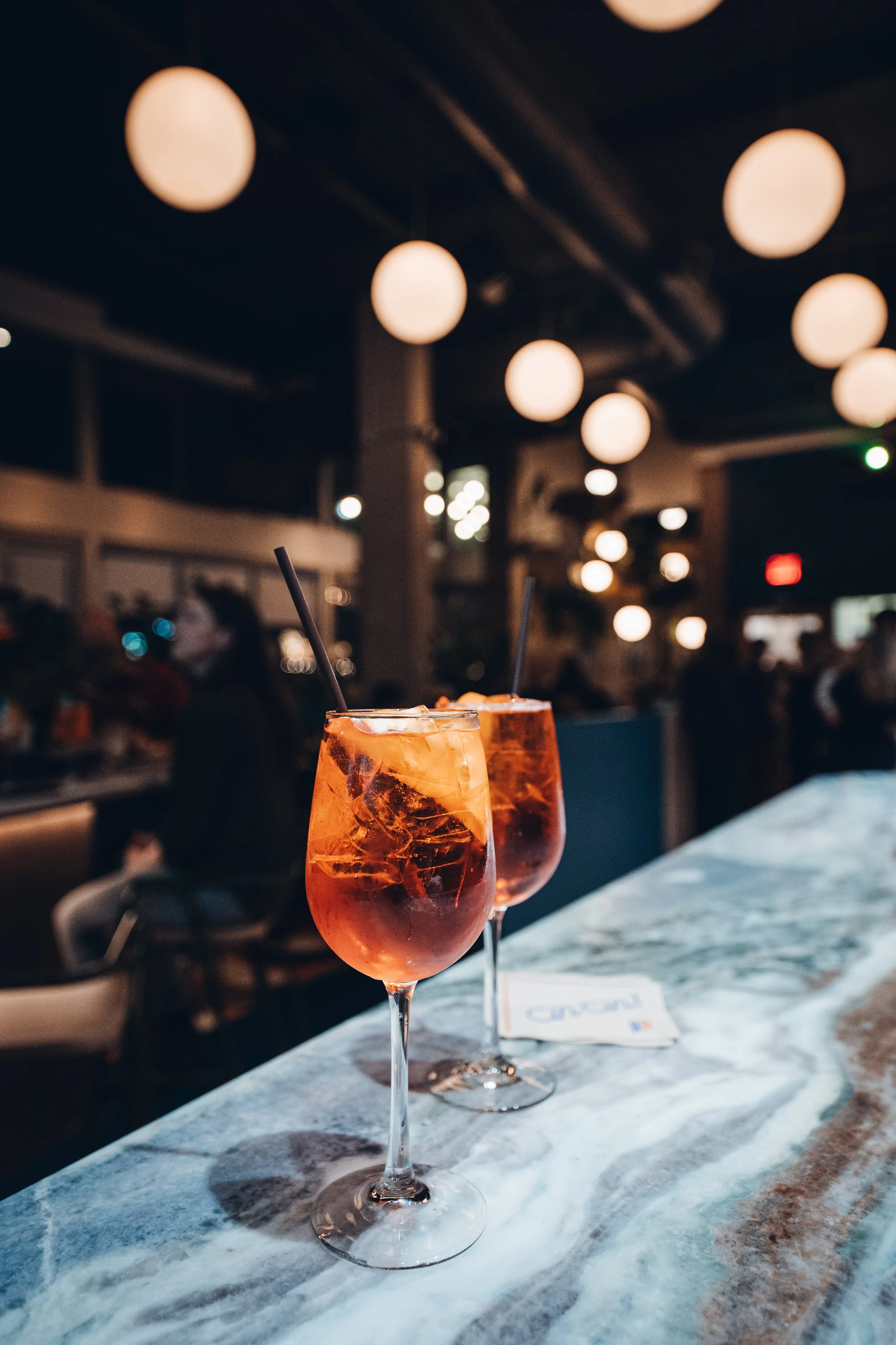 Two tall glasses filled with a spritz (orange-colored beverage and ice), garnished with black straws, on a marble bar counter in a dimly lit restaurant and warm overhead lighting.