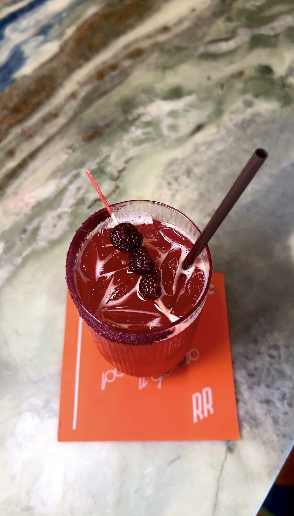 A tall glass of red cocktail with blackberries on top, ice cubes, and a brown straw, placed on an orange napkin on a marble table.