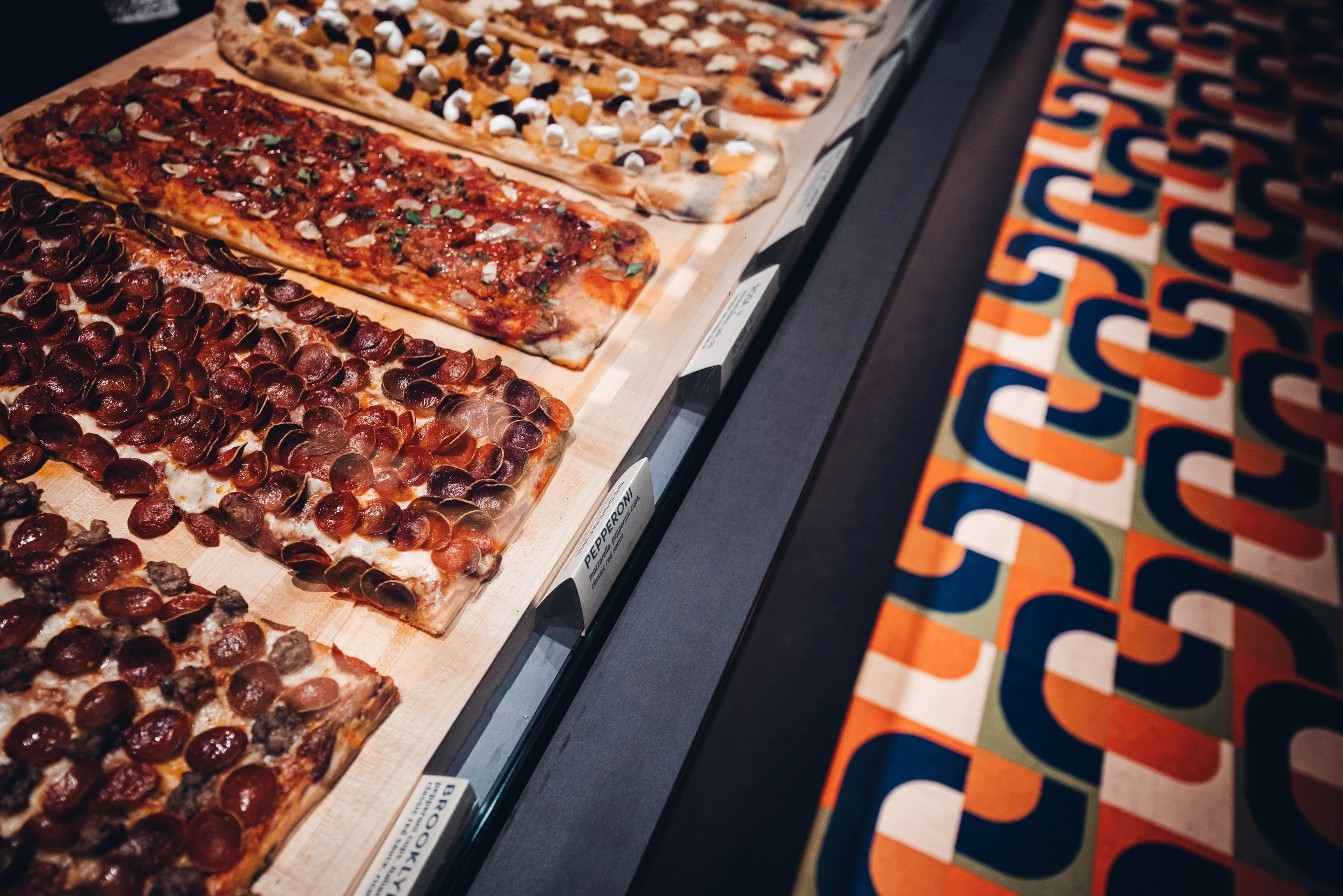 View of multiple rectangular pizzas with different toppings on display in a pizza case.