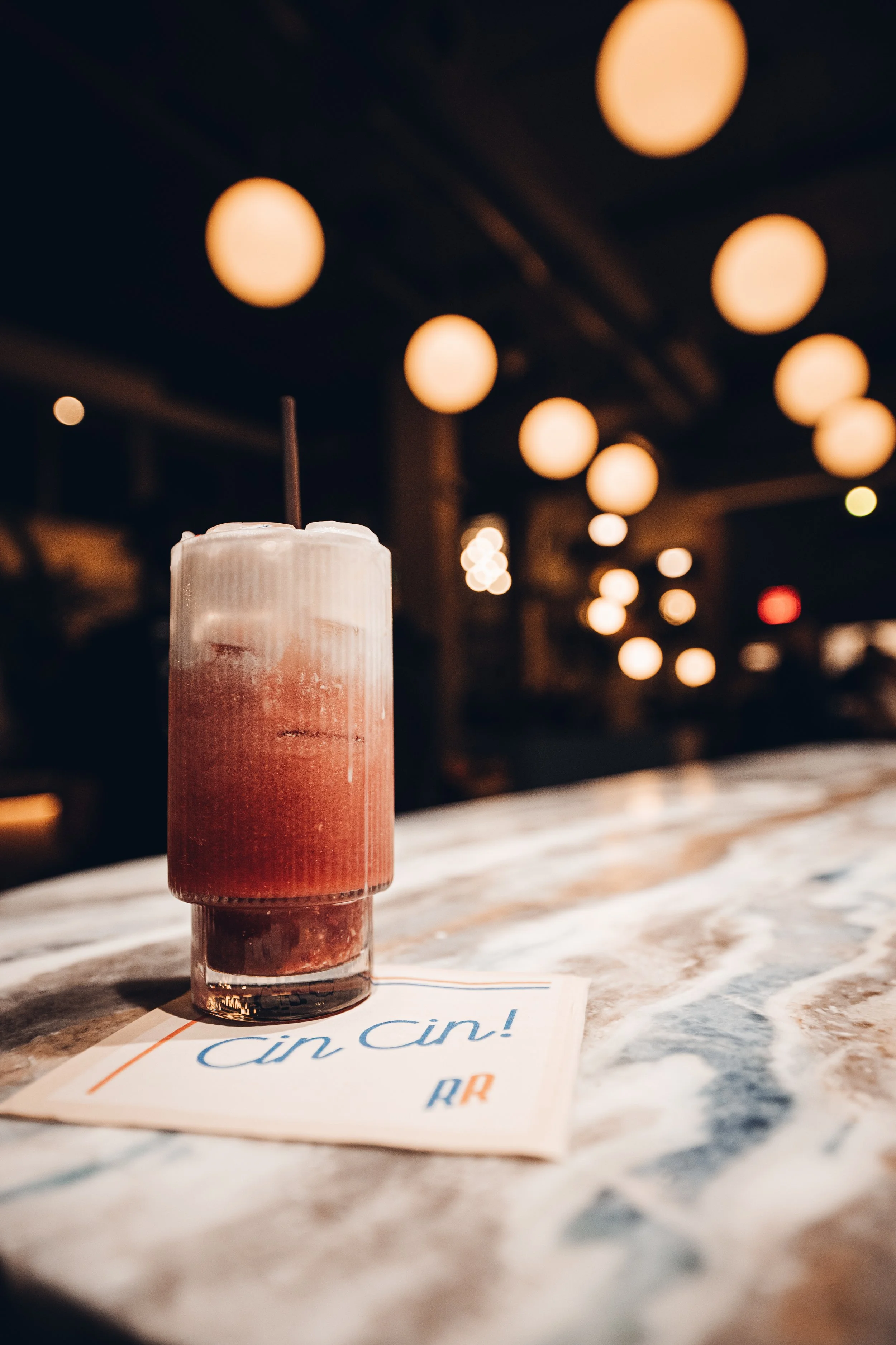 A glass of italian soda next to a handwritten note on a marble table, with warm bokeh lights in the background.