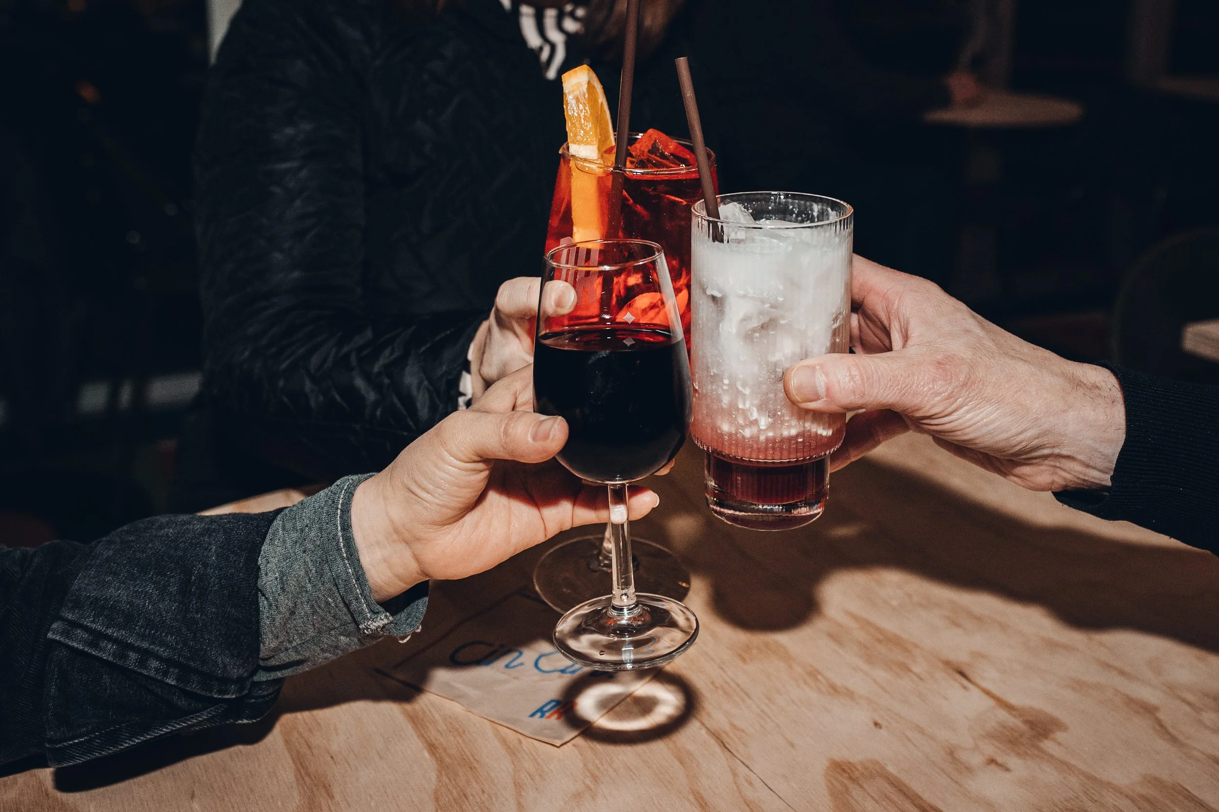 People clinking glasses with drinks, including red wine, a cocktail with an orange slice, and an italian soda, in a toast.