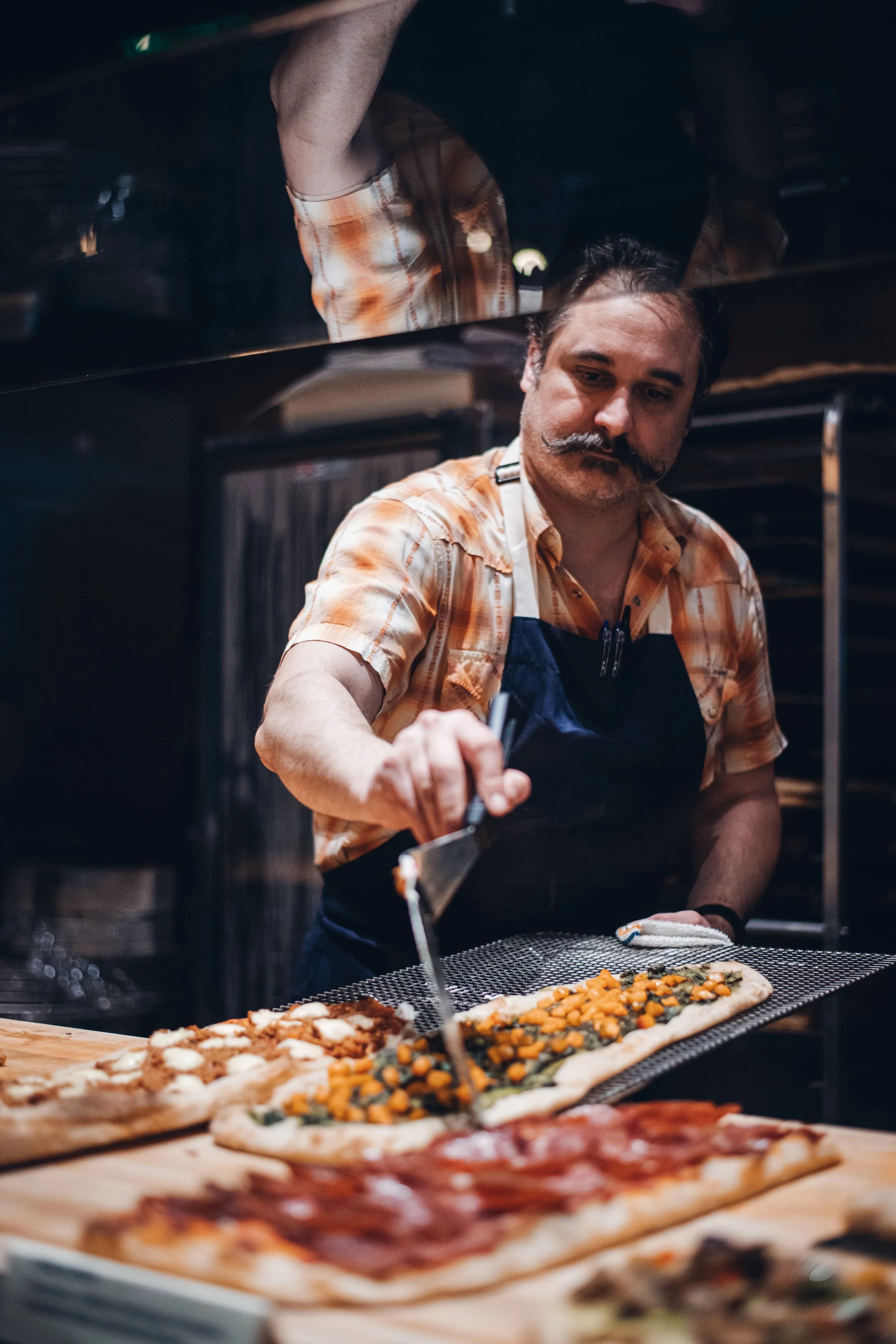 A man with a distinctive mustache in a plaid shirt and apron preparing pizza in a kitchen, with various toppings visible on the pizza.