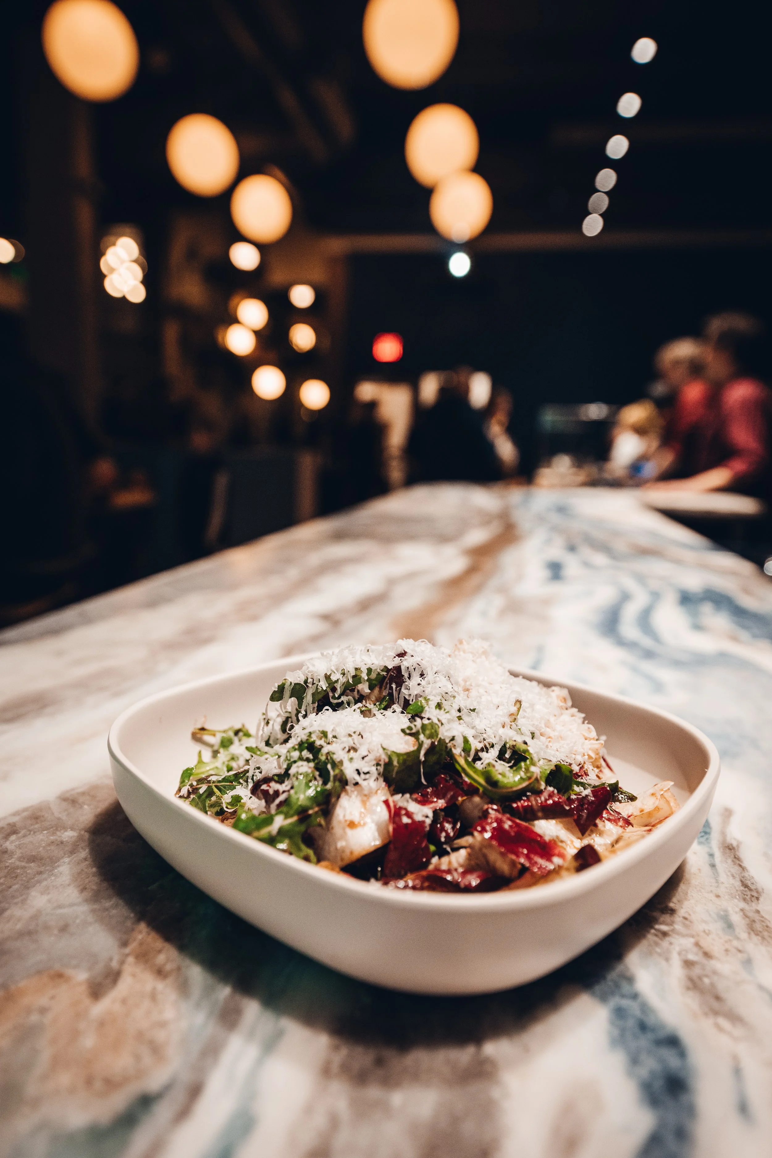 A bowl of salad with greens, shredded cheese, and chopped vegetables on a marble table in a dimly lit restaurant with blurred warm yellow lights and people in the background.