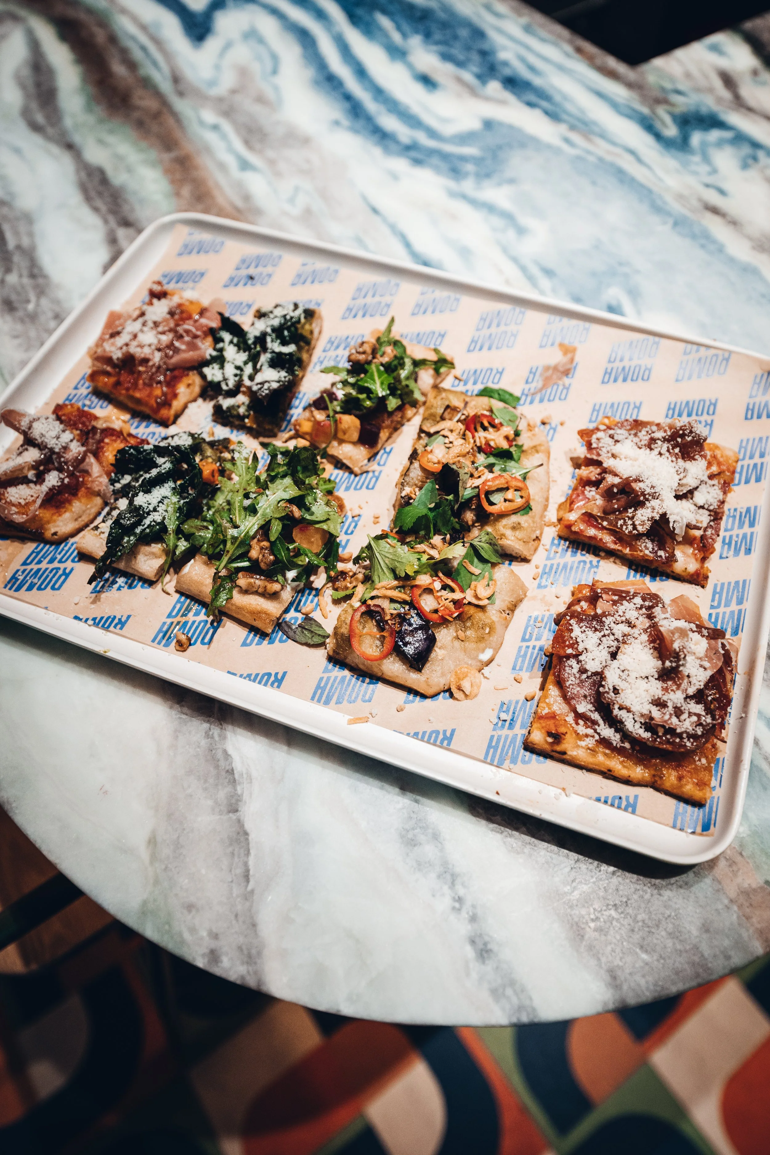 A white tray with several pieces of pizza topped with various vegetables, meats, and cheese, resting on a marble table. Each piece has a different topping and garnishes, such as herbs and sliced peppers.