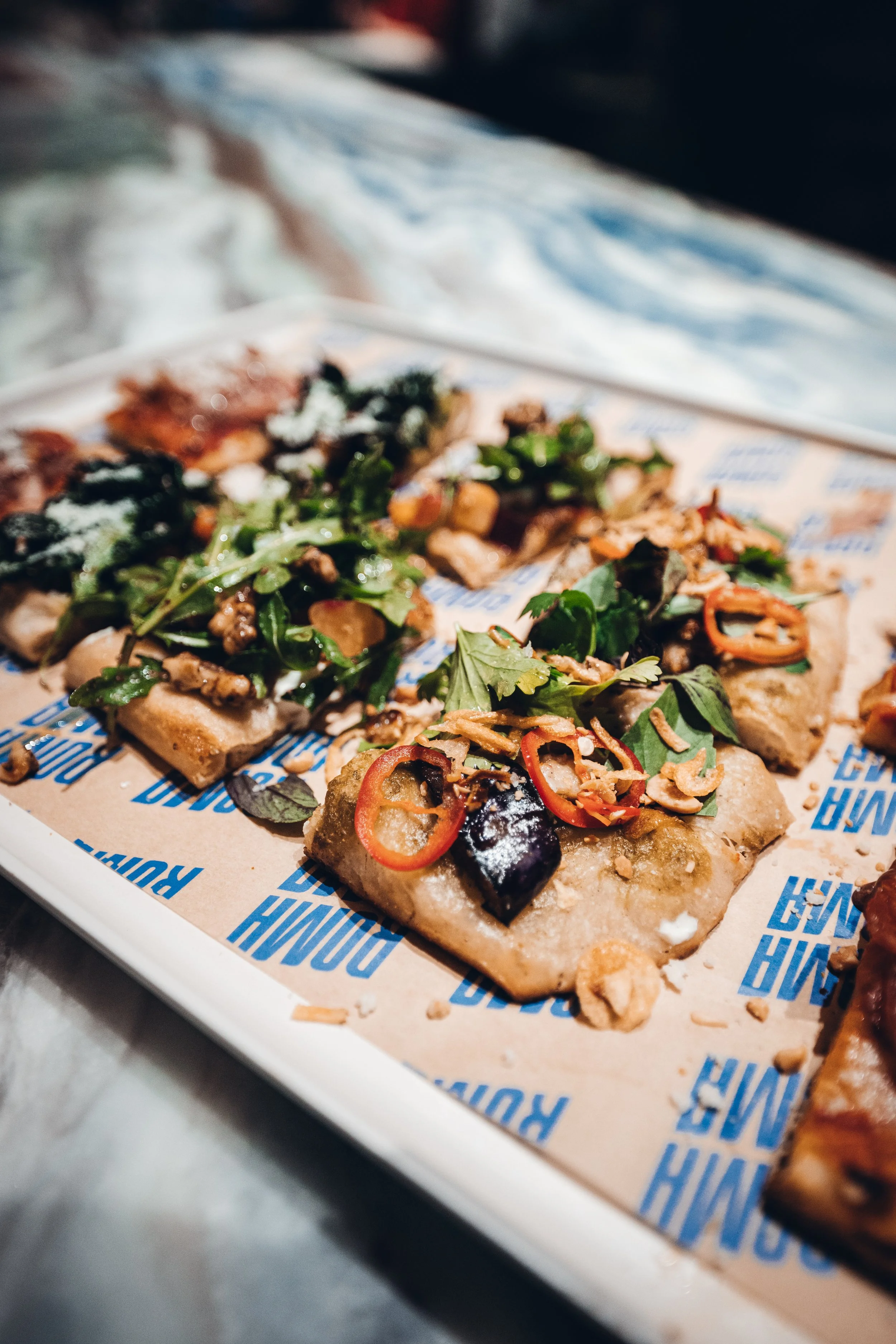 Close-up of assorted pizza slices on a tray, topped with vegetables, meats, and herbs, on branded paper lining.