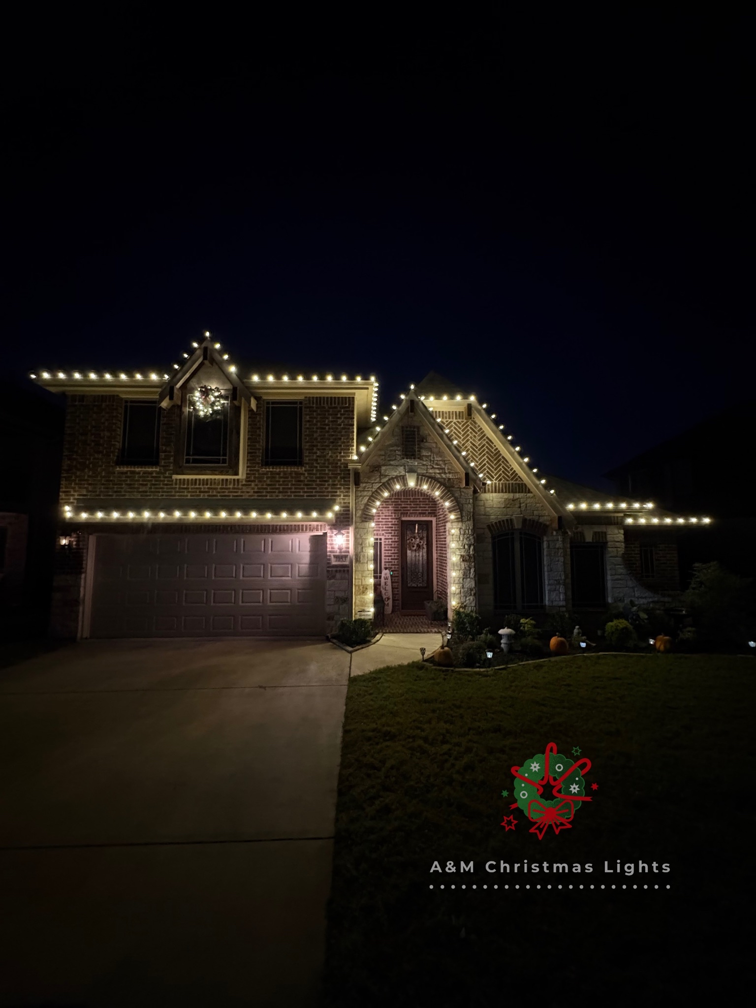 A house decorated with Christmas lights outlining the roof and windows, with Christmas decorations and pumpkins in the yard at night.
