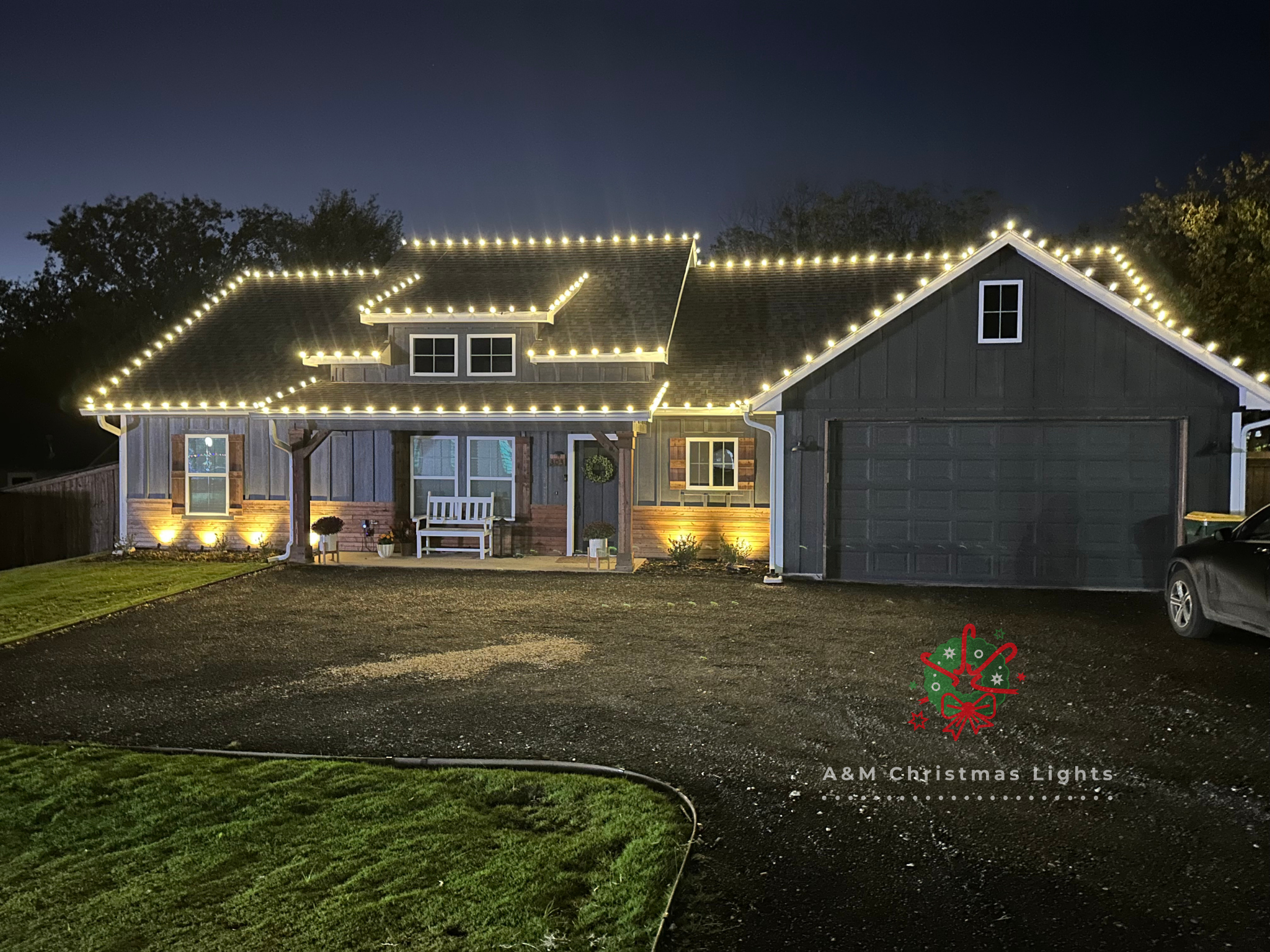 House decorated with white Christmas lights along the roofline, with a front porch with a bench, plants, and wreath, and a dark-colored garage door, on a night with dark sky and trees in the background.