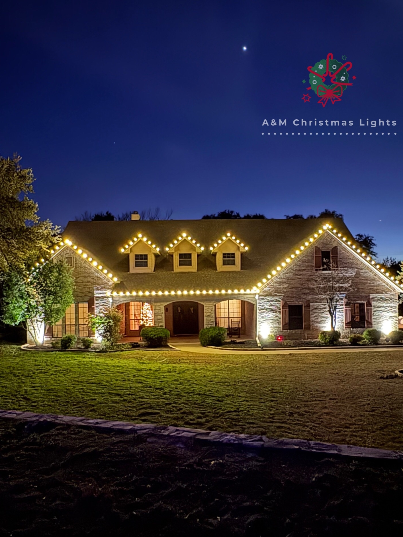 A house decorated with Christmas lights at night, with a dark blue sky and some stars visible. The house has a brick exterior and the yard is well-lit.