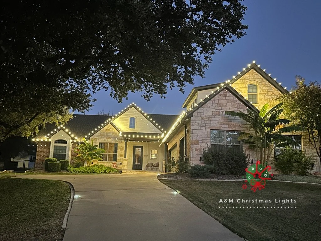 A house decorated with Christmas lights along the roofline, with a garden and trees in front, taken during dusk or early evening.