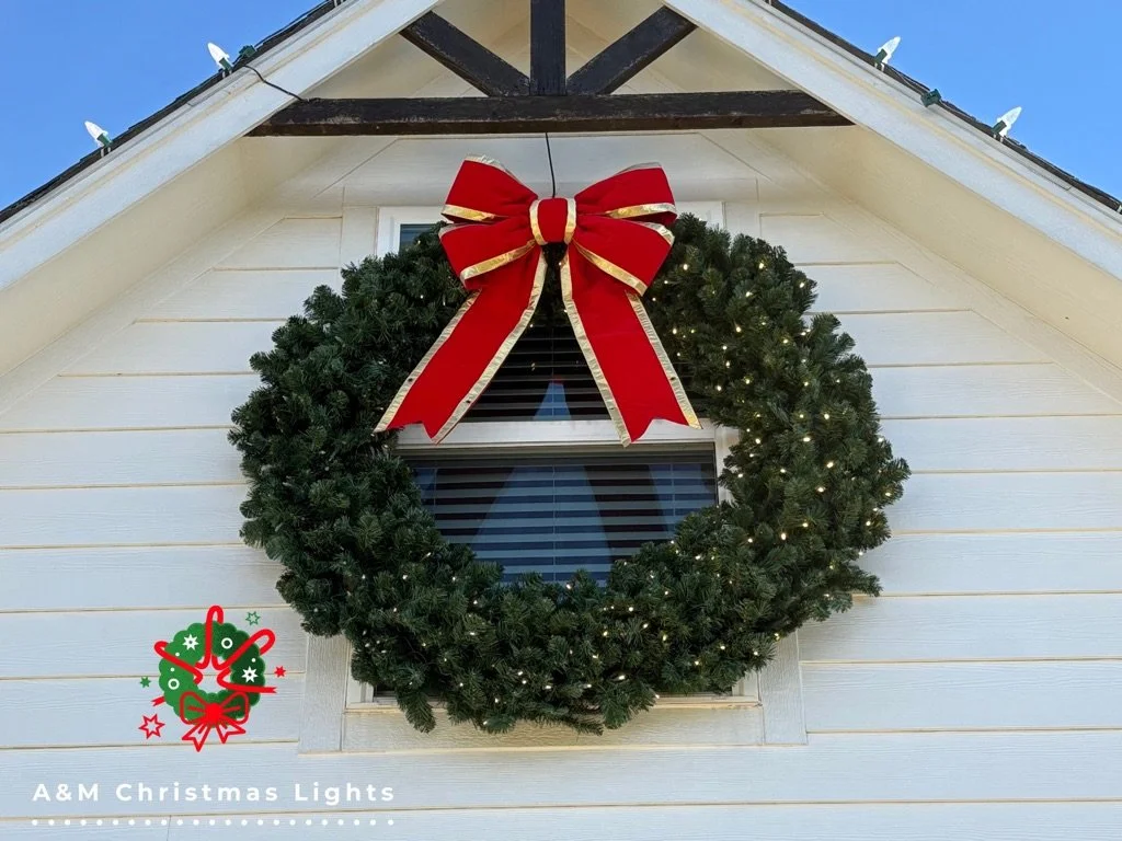 A Christmas wreath decorated with small white lights, hanging on the exterior wall of a house beneath the roof, with a large red bow on top.
