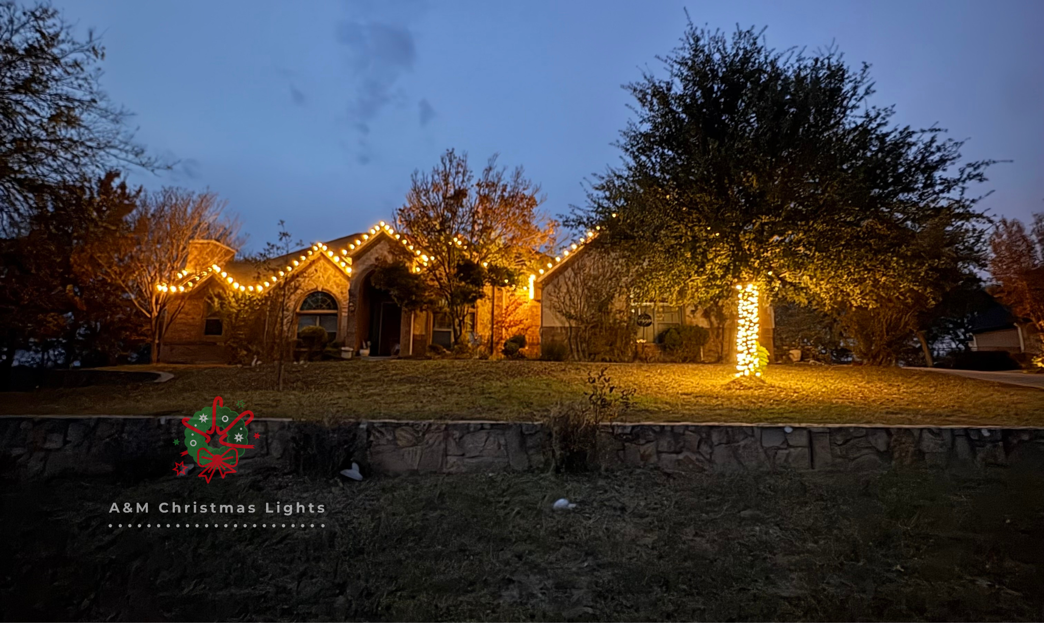 A house decorated with Christmas lights, including icicle lights along the roofline and a lit-up tree on the front yard, taken in the evening.