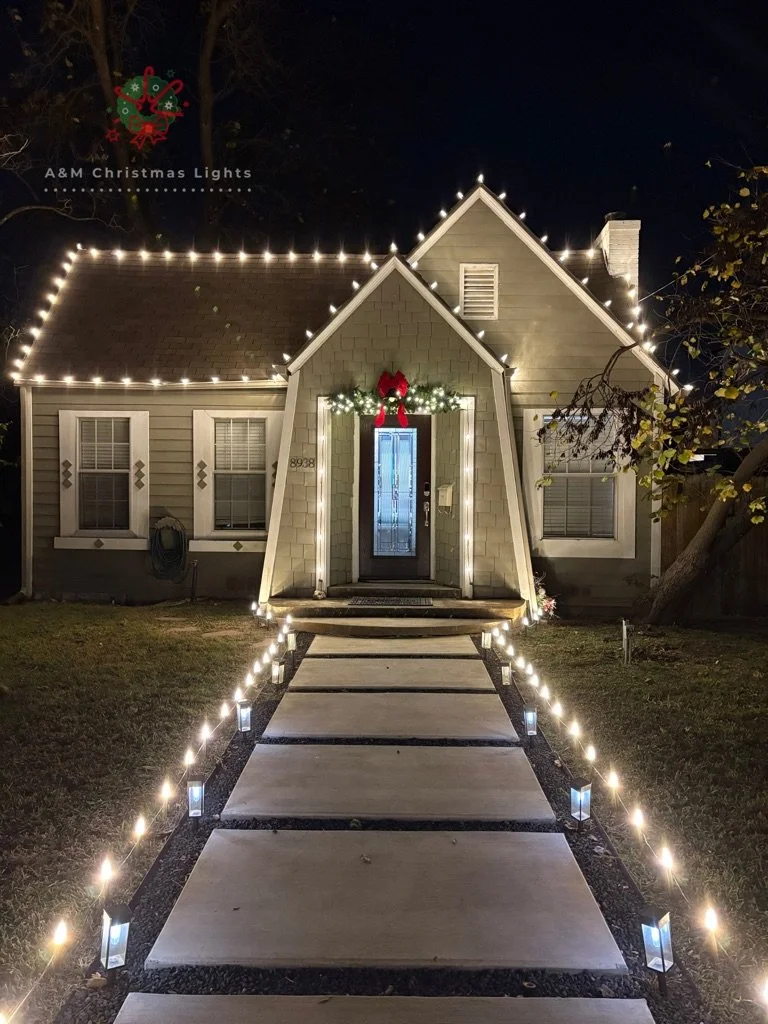 Nighttime exterior view of a house decorated for Christmas with white string lights outlining the roofline and front porch, a festive wreath with a red bow above the door, and a pathway lit with small ground lights leading to the entrance.