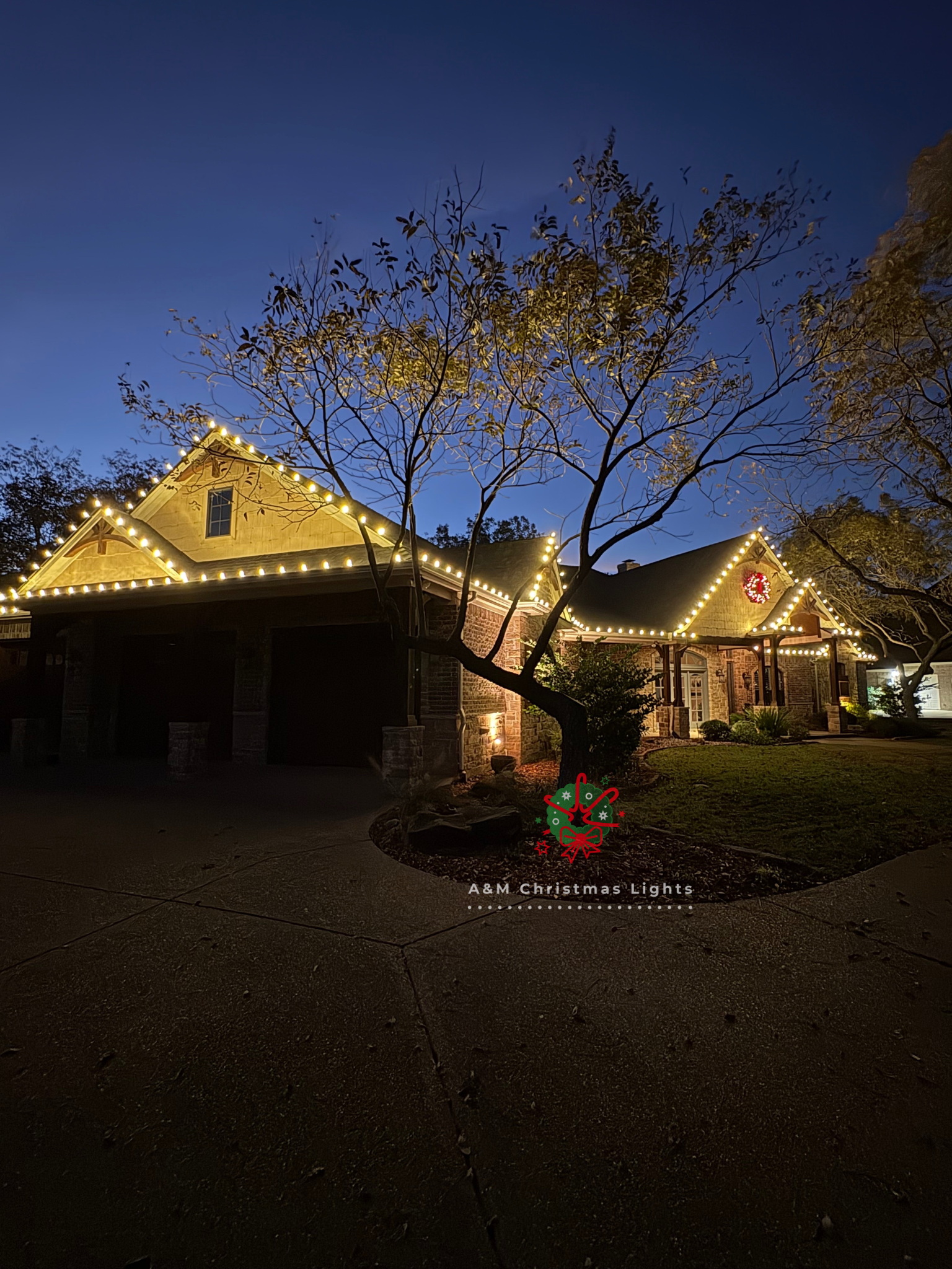 A house decorated with Christmas lights along the roofline, with a lit wreath on the front and trees in the yard, during the evening or night. The sky is dark blue.