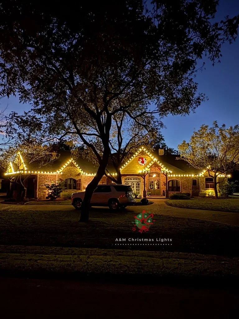 A house decorated with Christmas lights and a wreath on the front door, viewed in the evening or twilight, with trees and a vehicle in the front yard.
