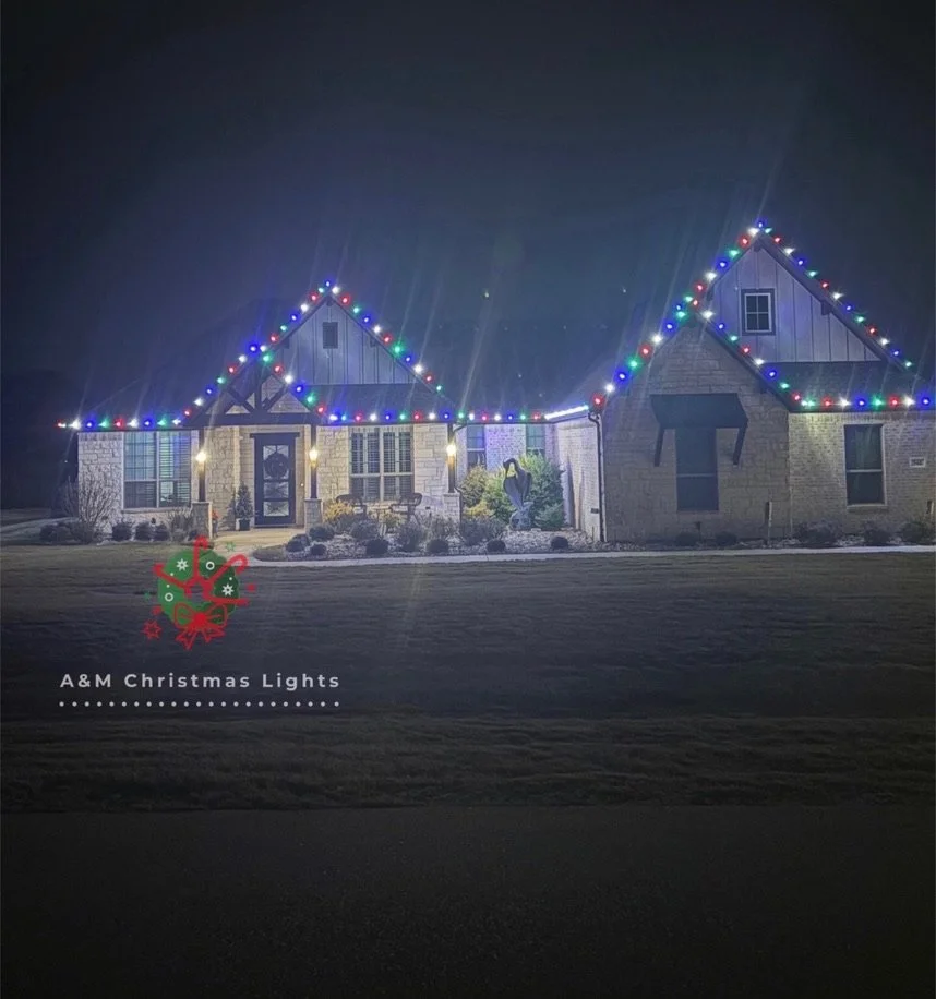 A house decorated with colorful Christmas lights along the roofline at night. Front yard has bushes and a small tree. There is a text overlay with a Christmas ornament graphic and the words "A & M Christmas Lights."