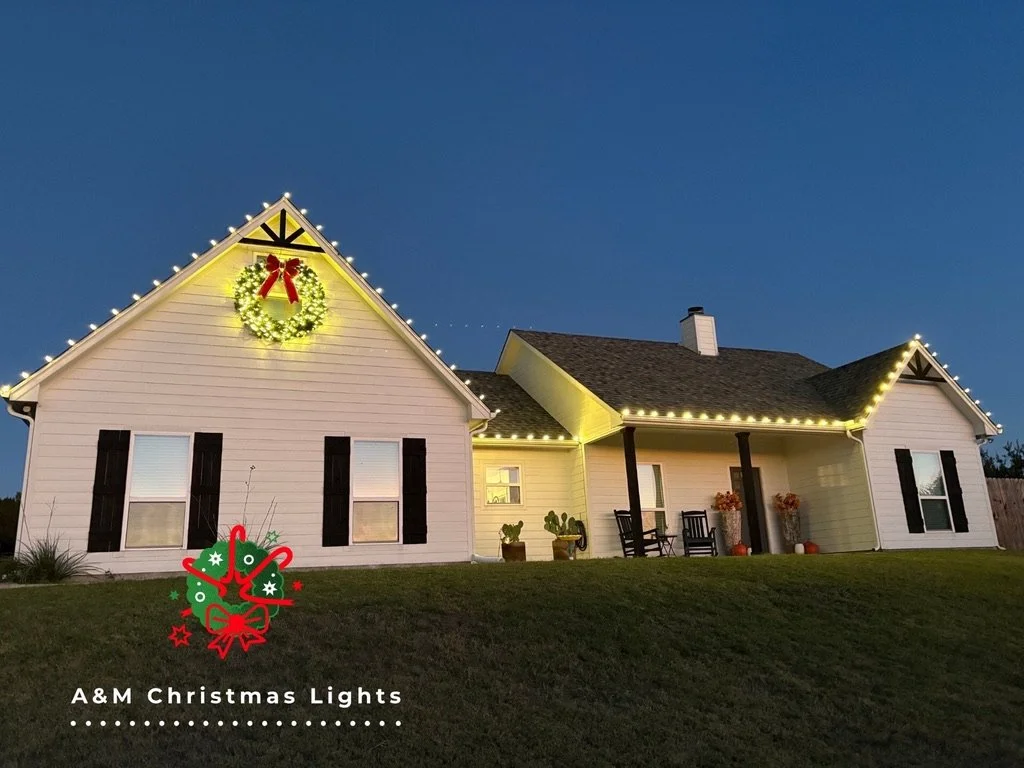 A house decorated with Christmas lights and ornaments, including a large festive wreath with a red bow on the gable, and a string of lights outlining the roof. There is a holiday graphic with a decorated Christmas tree, ornaments, and the text "A&M C