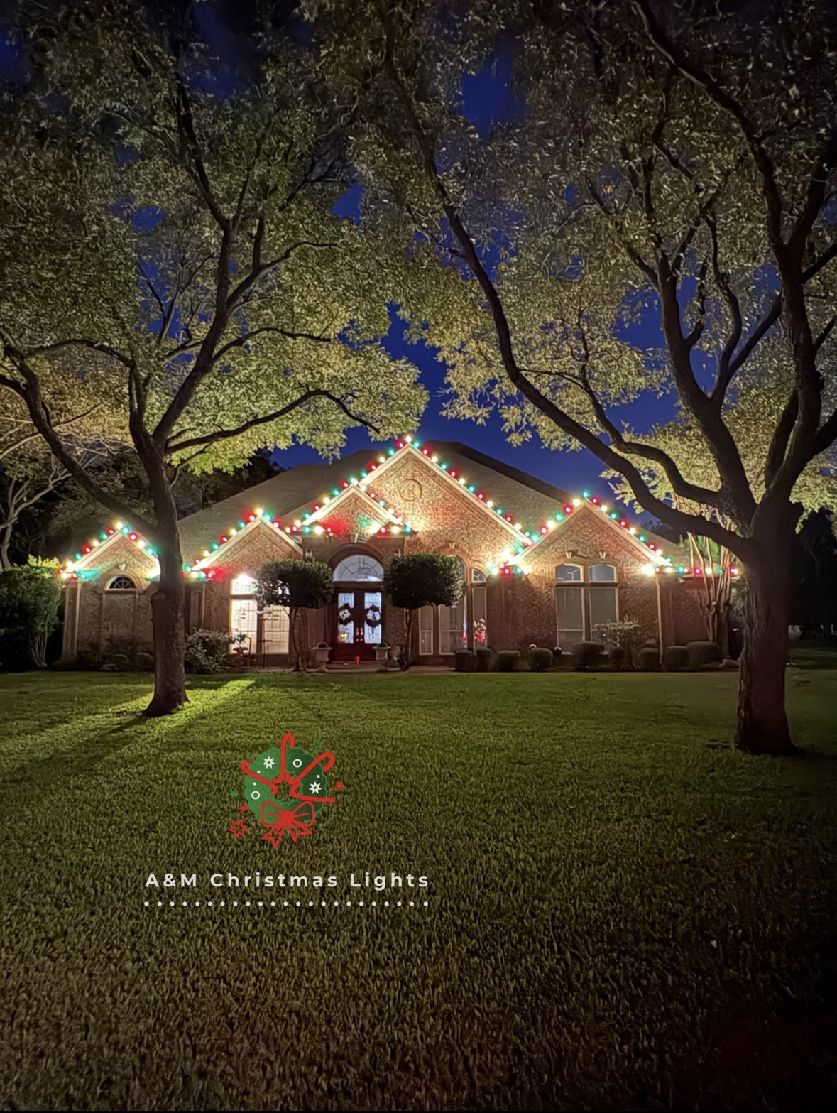 A house decorated with multicolored Christmas lights along the roofline, with trees and a well-maintained lawn in the foreground during the nighttime.