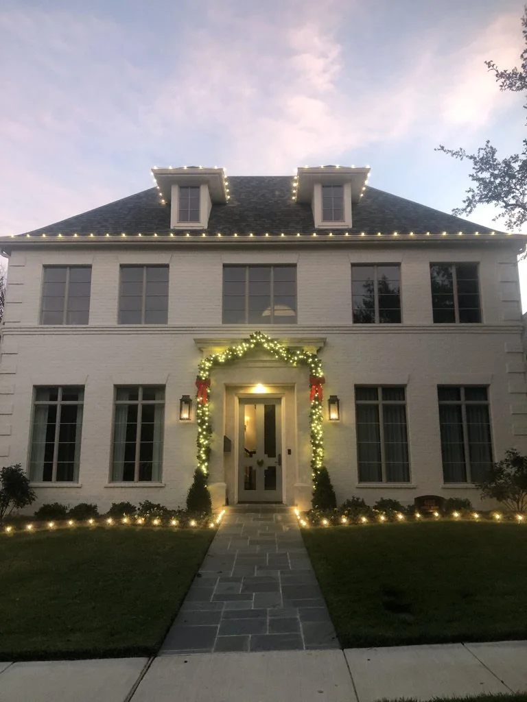 A large white house decorated with Christmas lights and garlands. Lights outline the roof and dormer windows, and the front door is adorned with Christmas greenery. The house has a stone pathway leading to the front door, with a well-manicured lawn o