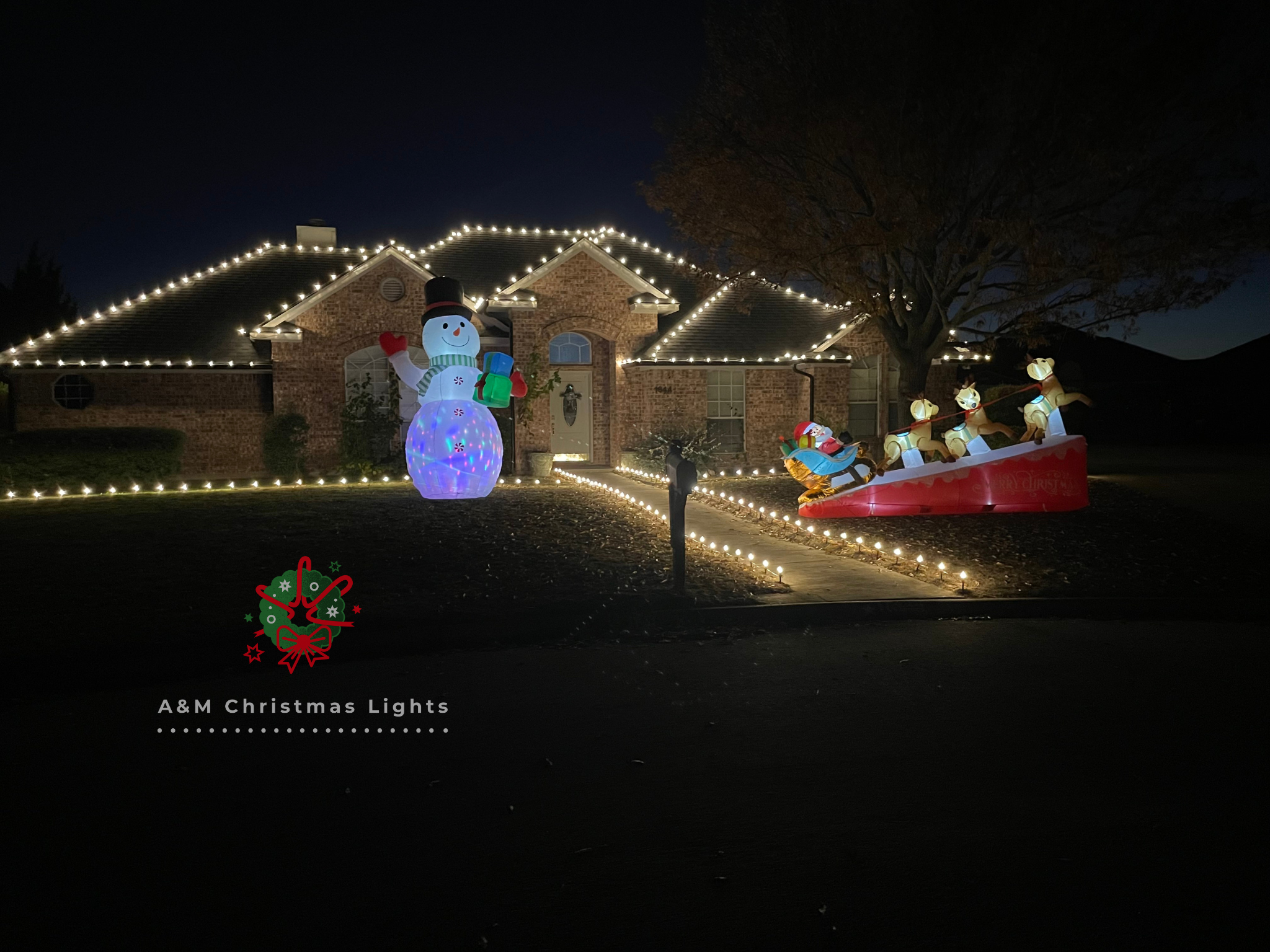 Nighttime Christmas display in front of a brick house with string lights on the roof. Features a large illuminated snowman and a Santa's sleigh with four reindeer.