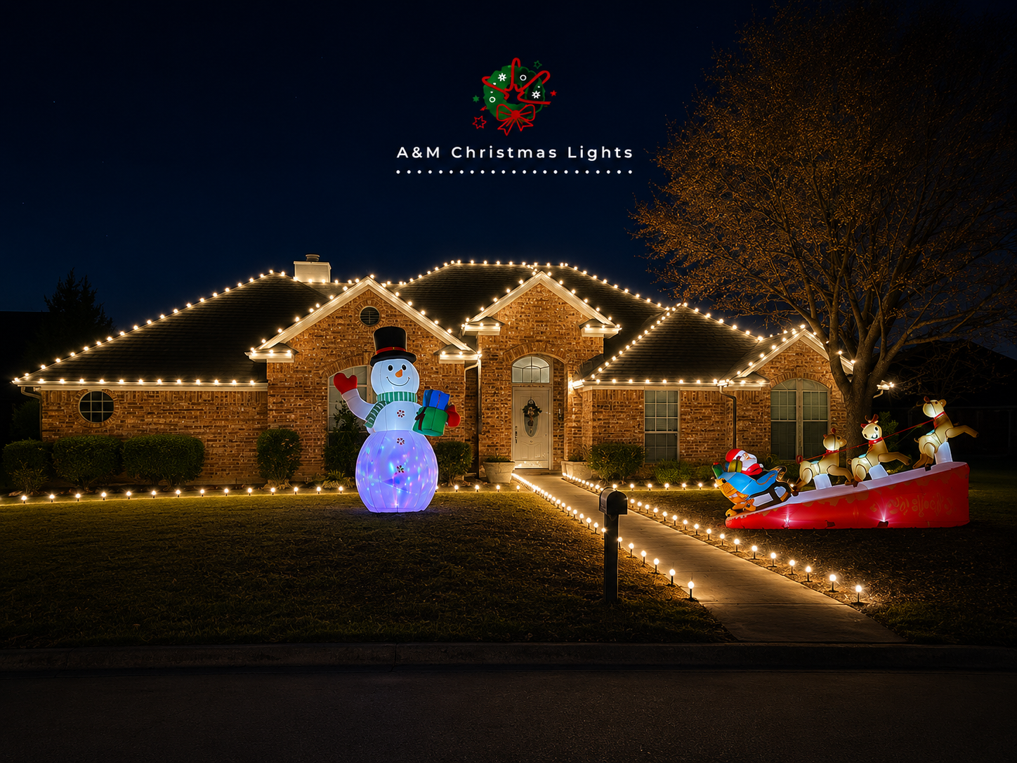 A brick house decorated with white Christmas lights outlining the roof. In front, there is an inflatable snowman holding gifts, and a toy sleigh with four reindeer decorations. The scene is illuminated at night and appears festive for Christmas.