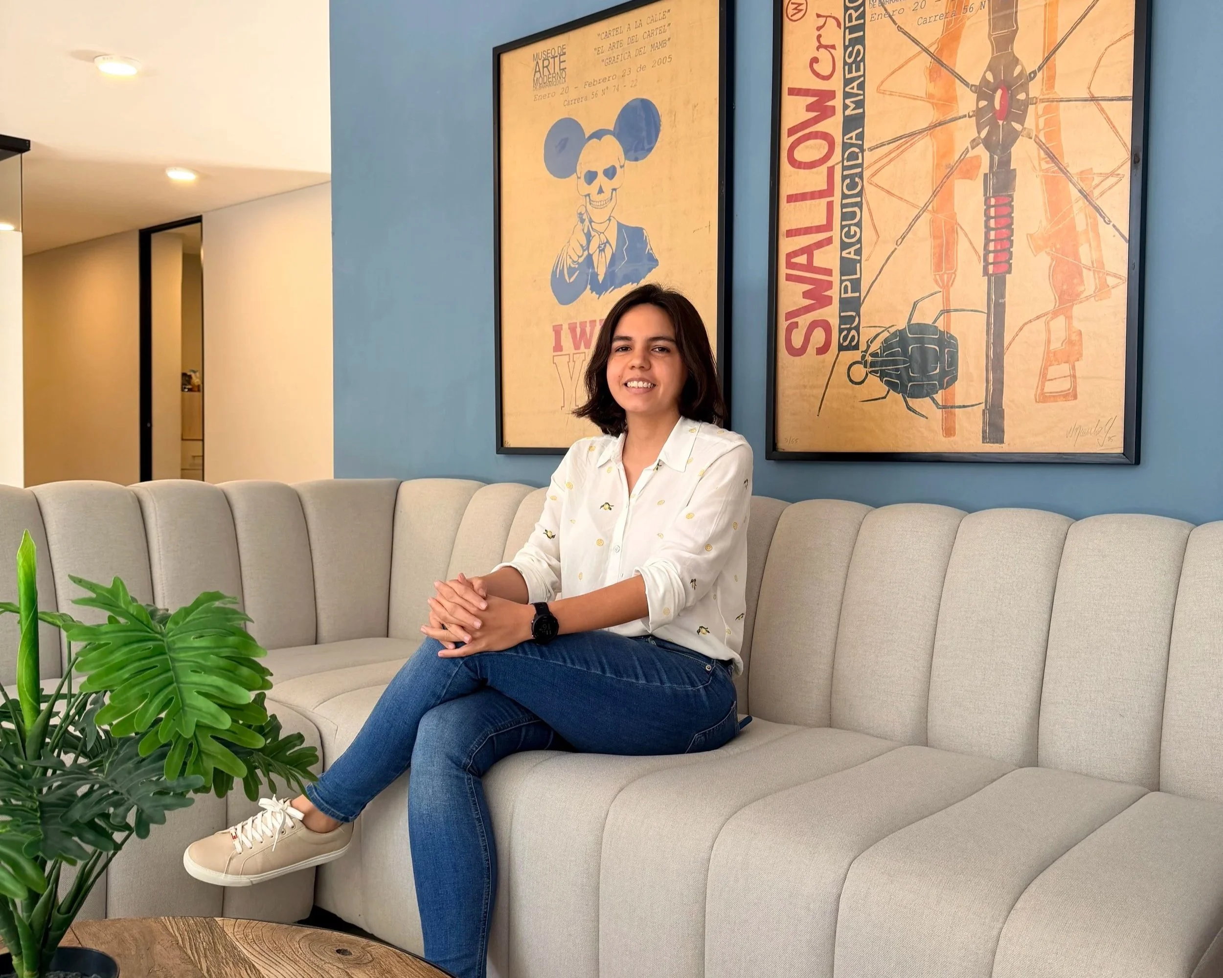 Una joven sentada en un sofá beige, con fondo de paredes azules y cuadros con arte gráfico, rodeada de plantas verdes en un espacio moderno y bien iluminado.