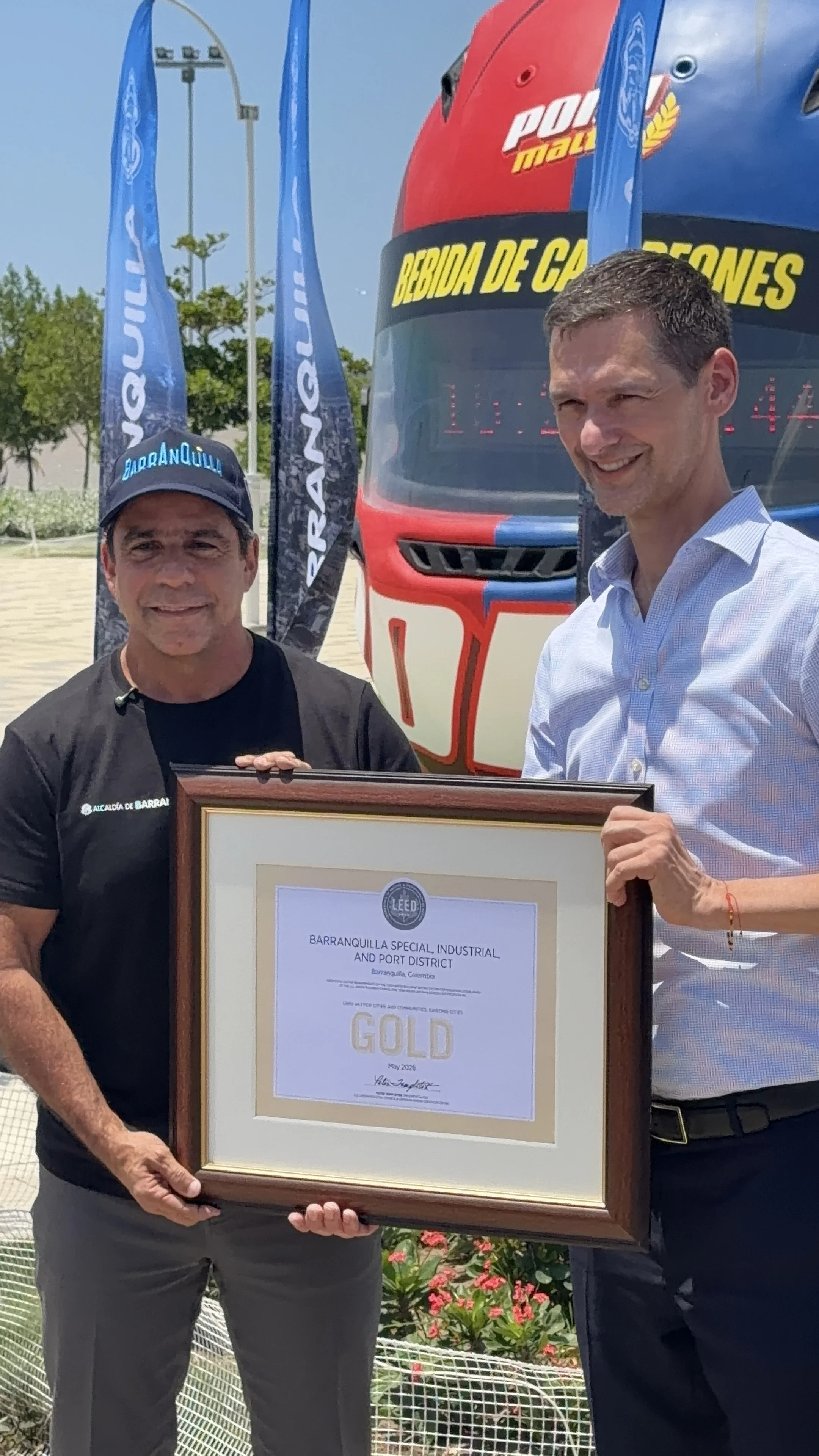 Dos hombres sosteniendo un marco con una certificación, uno viste una camiseta negra con la leyenda 'ALCALDÍA DE BARRANQUILLA' y gorra azul, el otro con camisa de manga larga azul y sonrisa. Detrás hay un autobús con la inscripción 'BEBIDA DE CARBONE