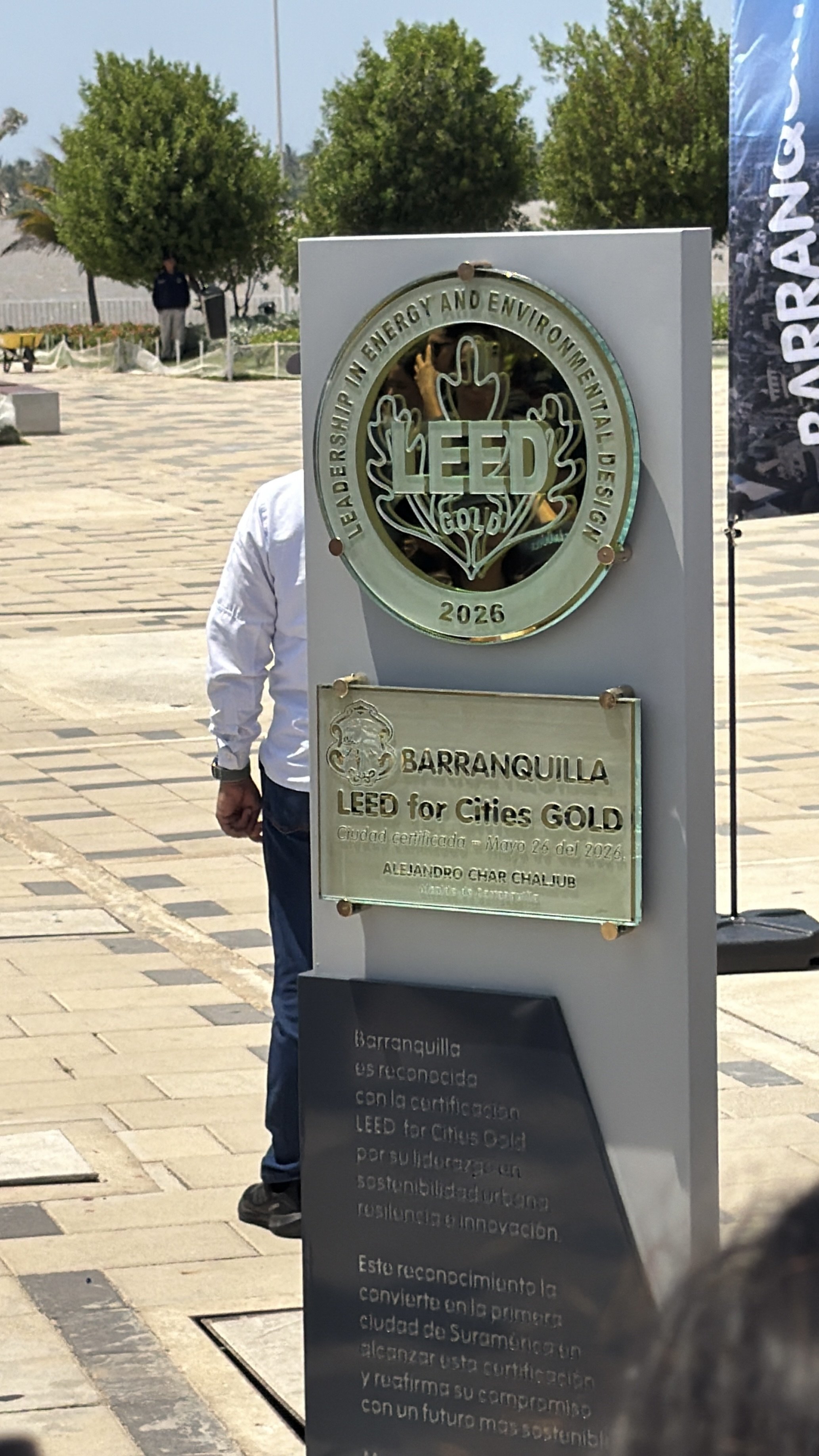  cartel dorado y plateado con inscripciones sobre certificación LEED en la ciudad de Barranquilla, con árboles de fondo y una persona parcialmente visible junto a él.