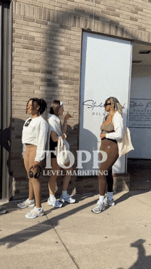 Four women standing outdoors near a brick wall and a glass door, engaged in conversation, with shadows cast on the ground.