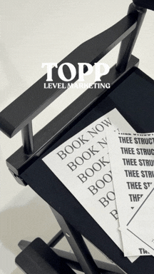 A black desk with a cardboard cutout that reads "TOPP LEVEL MARKETING" and several printed sheets of paper with the words "BOOK NOW" on them.