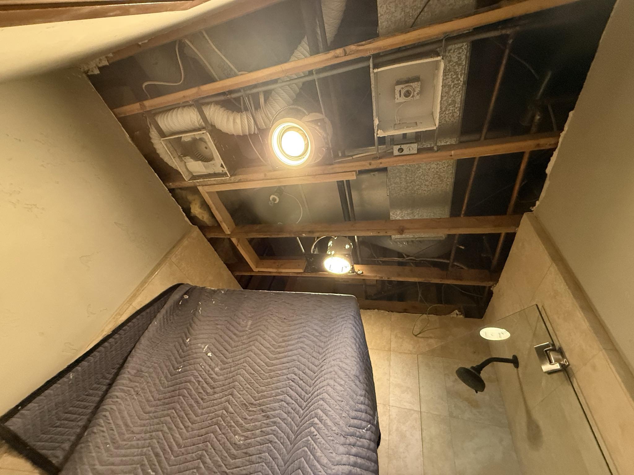 Unfinished bathroom with exposed ceiling framing and ductwork, a bed with a dark quilted cover, and a shower area with a black showerhead.