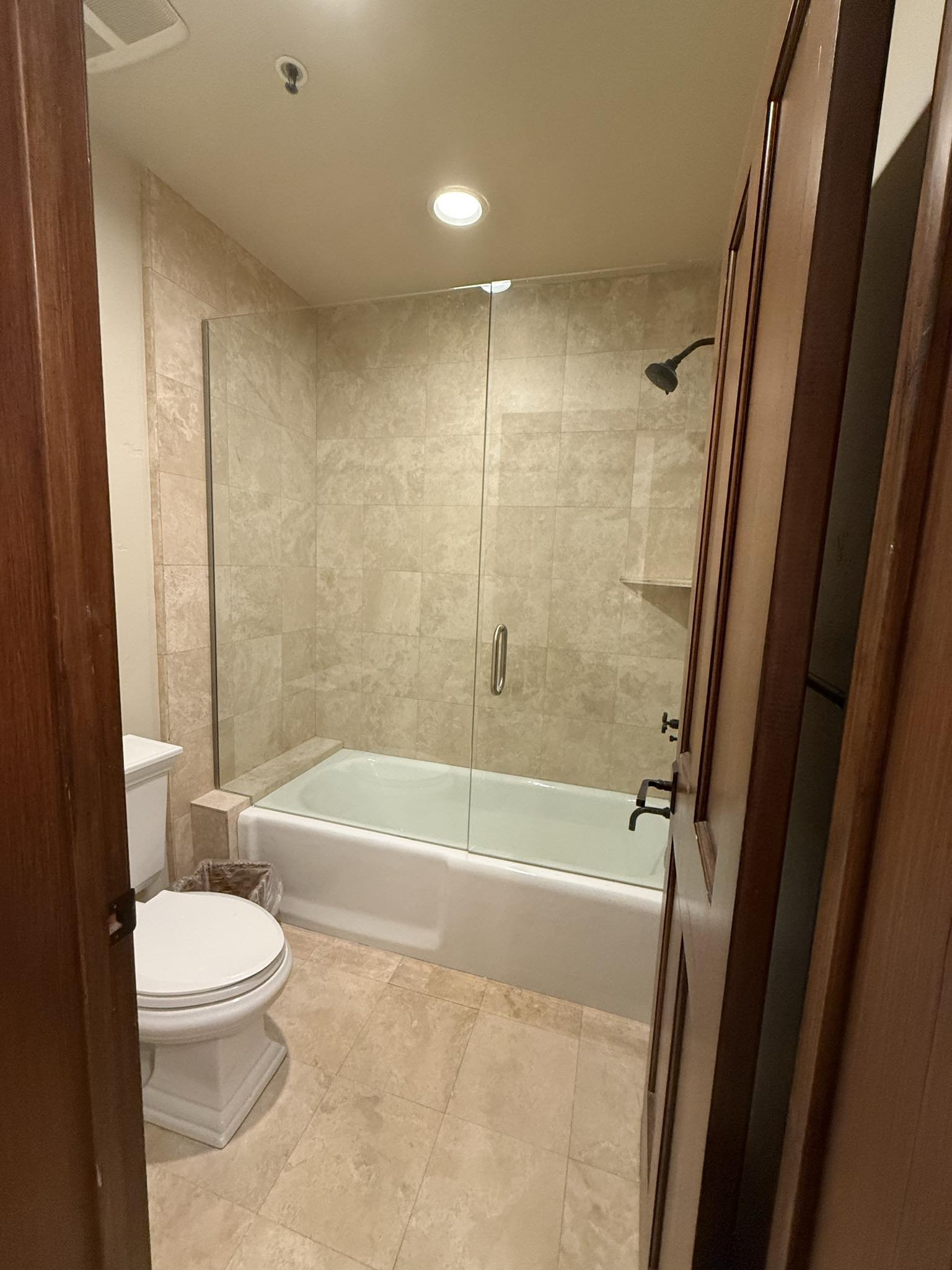 Bathroom with a bathtub, clear glass shower door, beige tiles, and a toilet next to the bathtub, with a small wastebasket beside it.